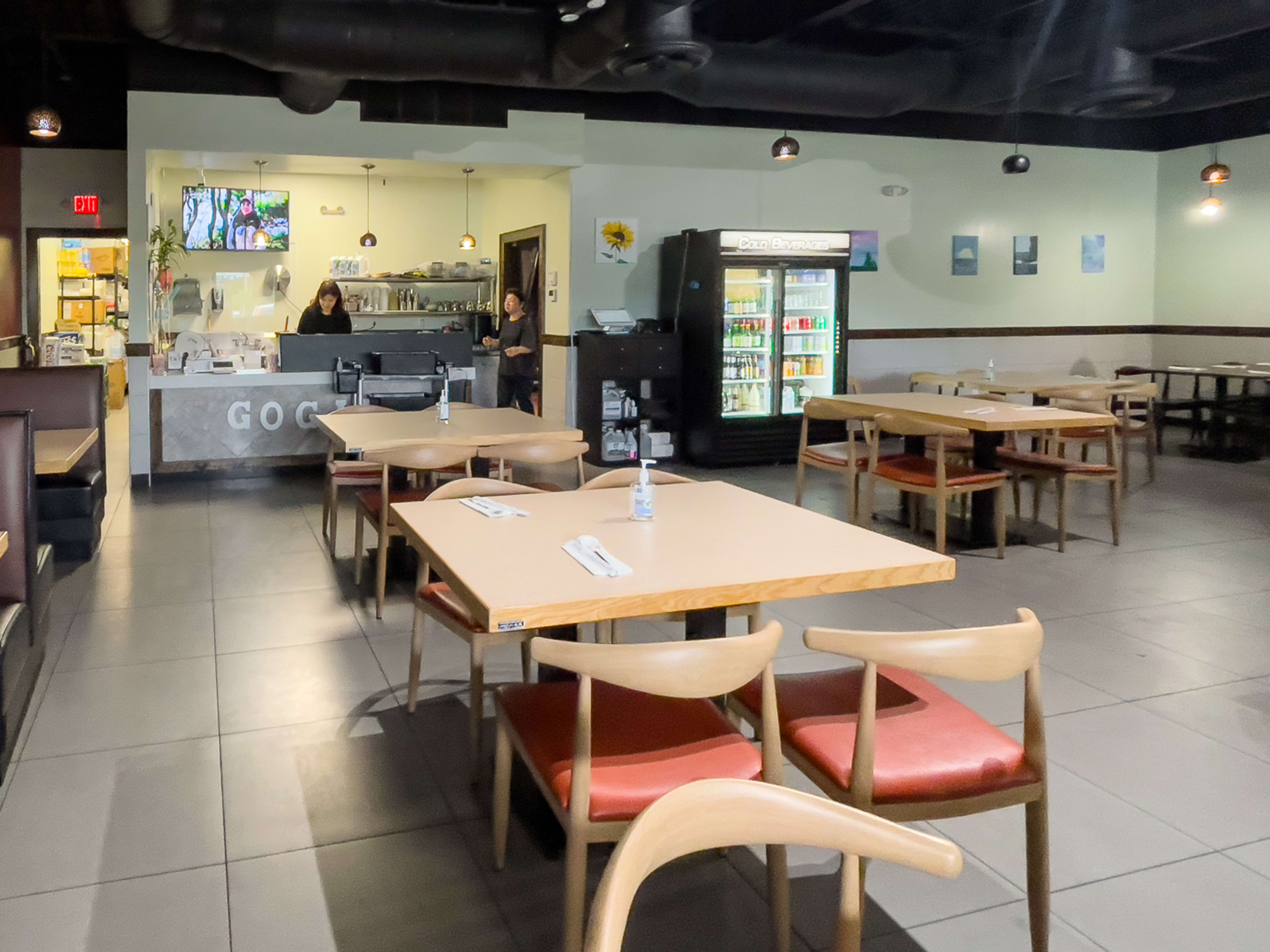 Restaurant interior with ordering counter, fridge of bottled drinks, wooden tables and chairs, and booths.