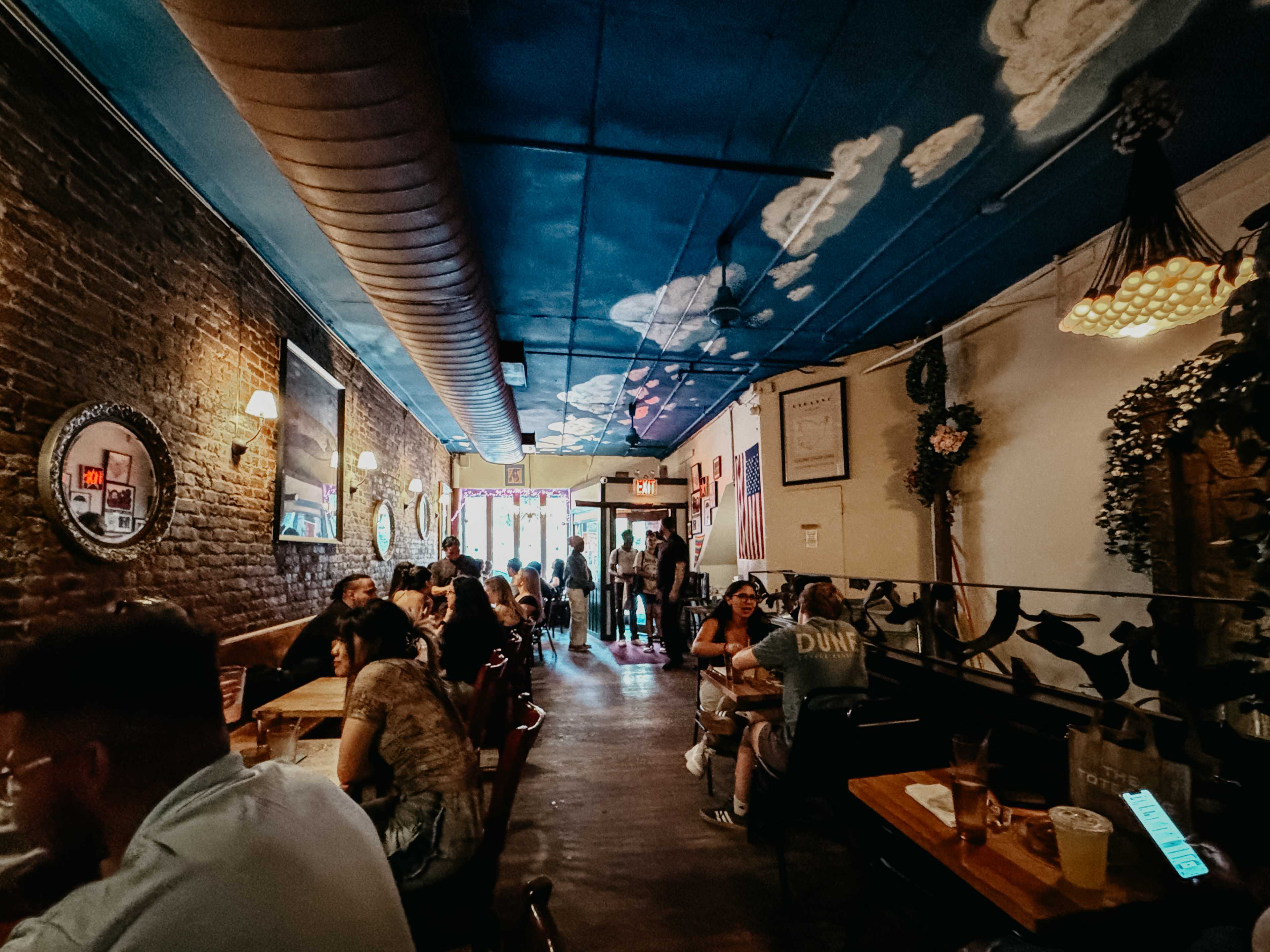 A crowded restaurant with little wooden tables and clouds on the ceiling.