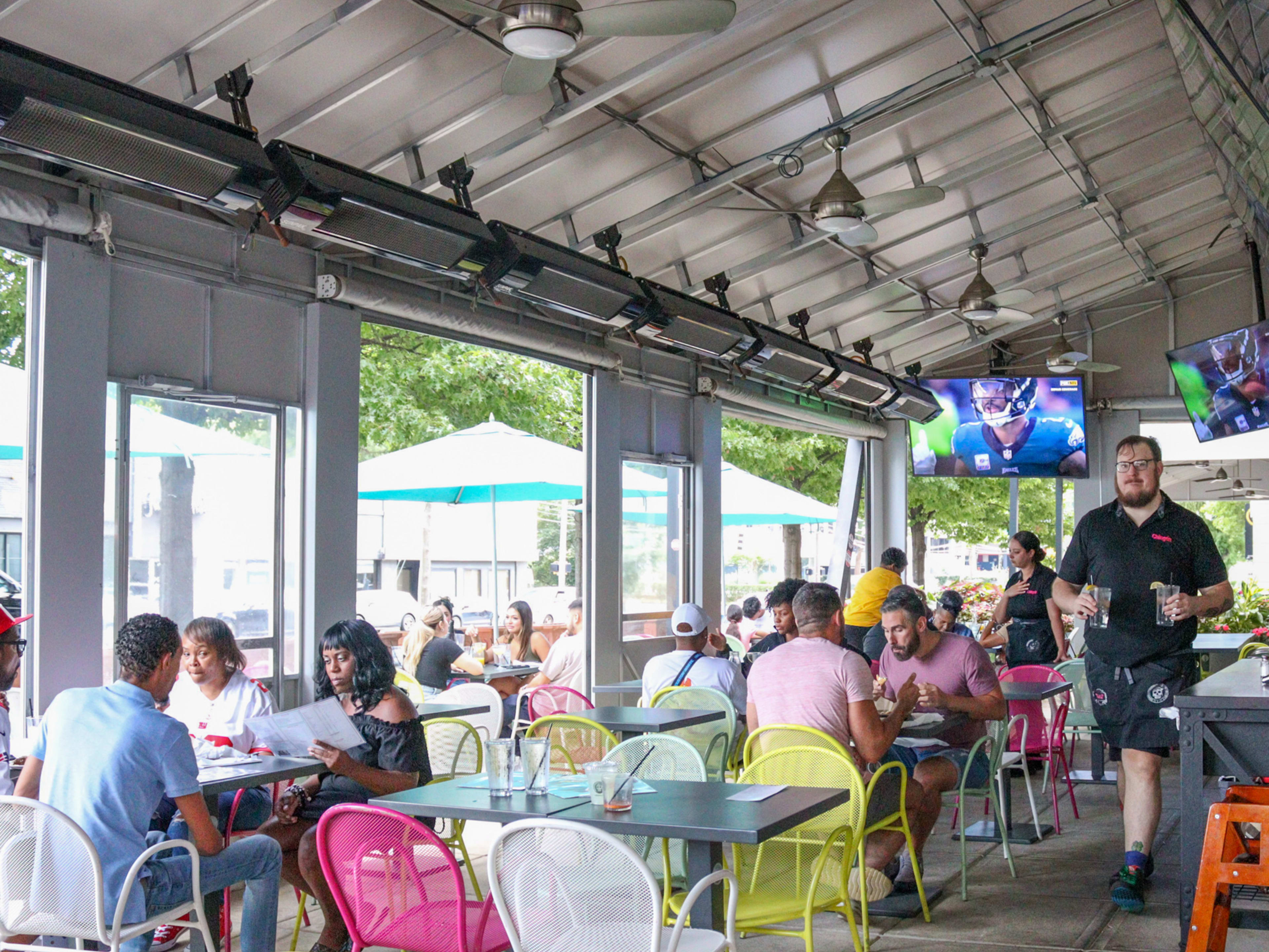 Covered heated patio with people dining at tables and mounted tvs playing sports.