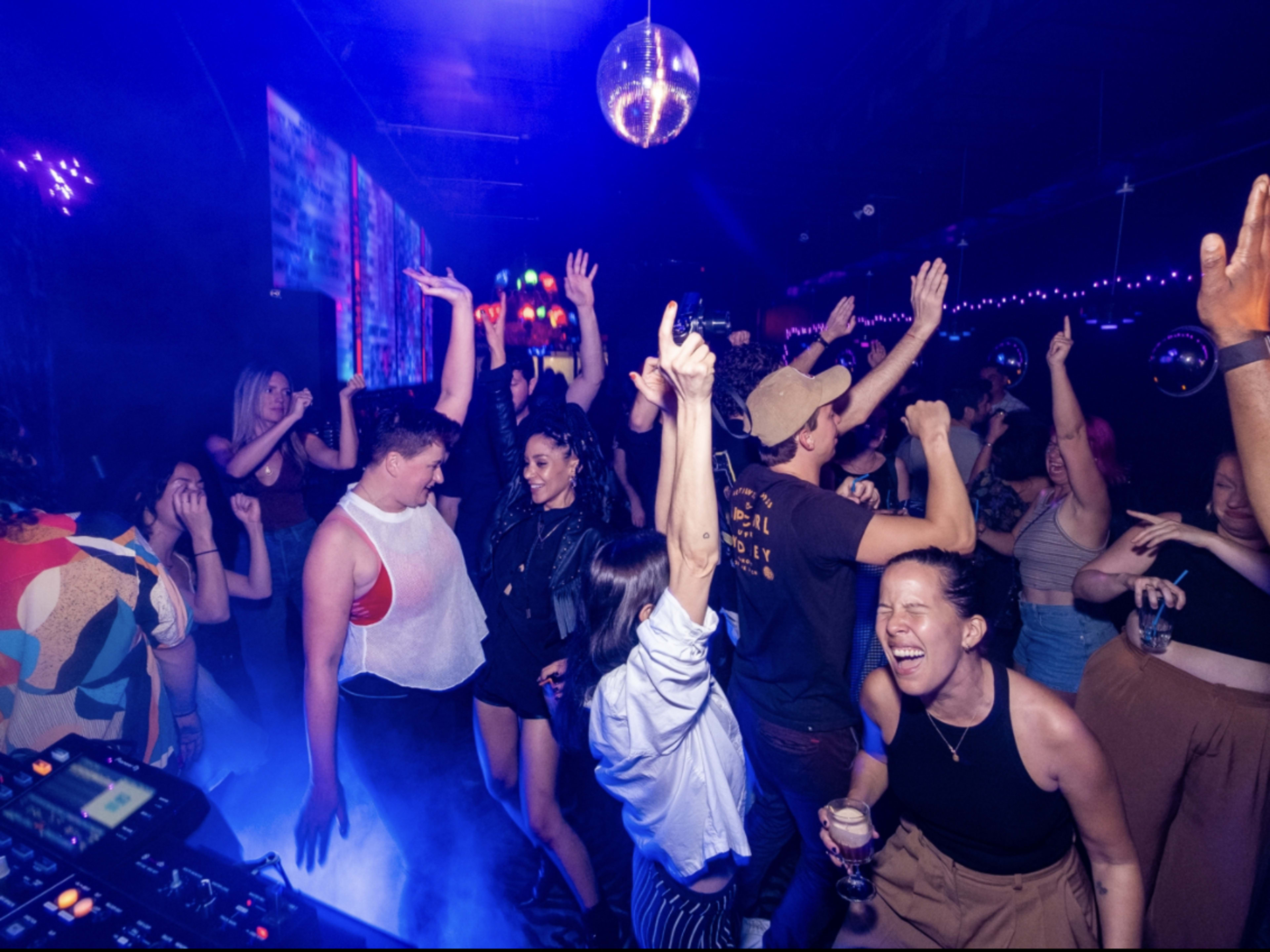 A crowd of people dancing inside Danger Danger bar.  There is a disco ball and blue and purple lighting. A DJ booth is set up in front of the dance floor.