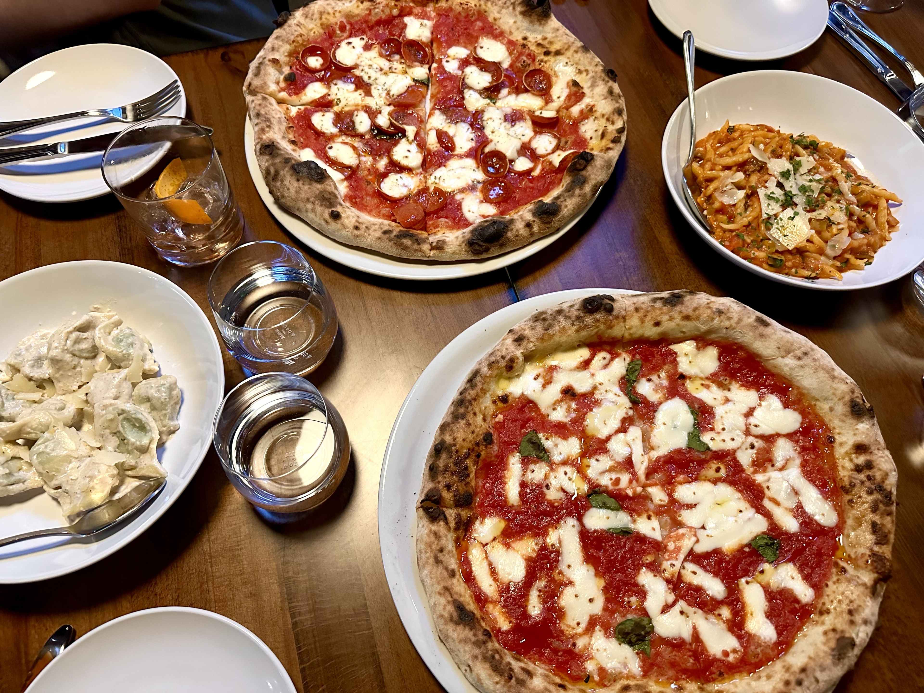 Two Neopolitan pizza pies and two bowls of pasta at a wooden table