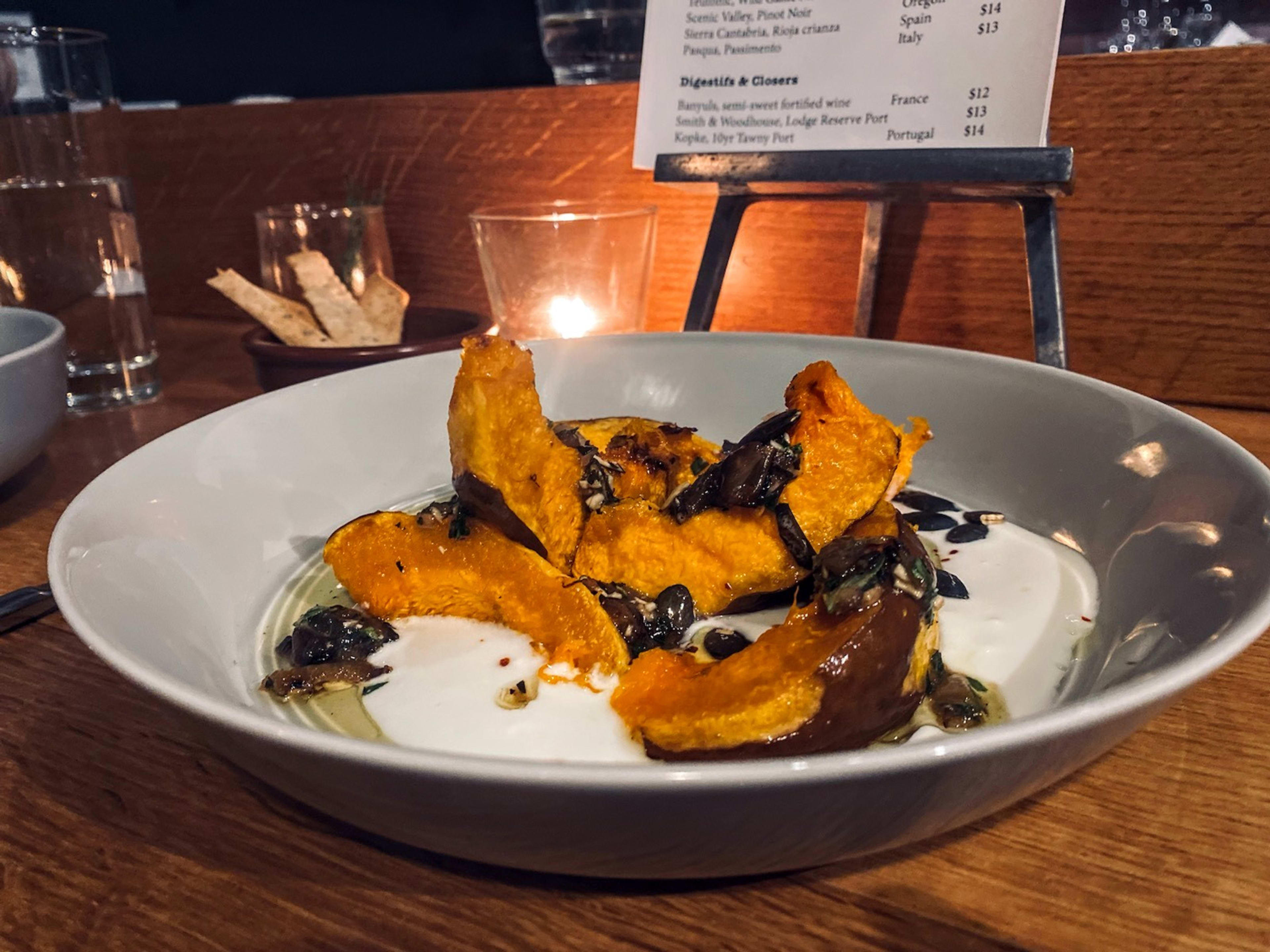 A candlelit table with a small bowl of crackers and a larger bowl containing roasted squash slices resting on a white sauce.