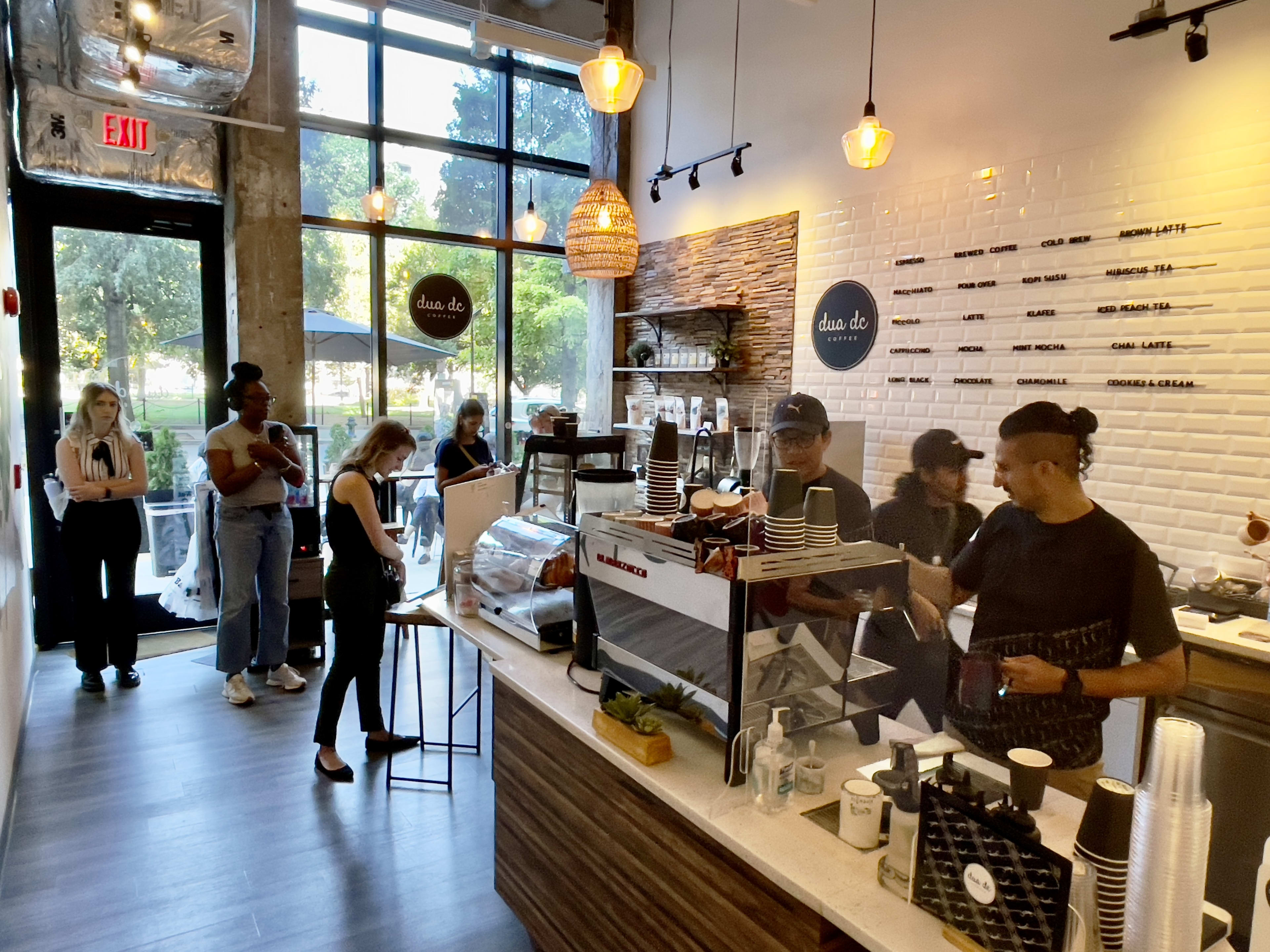 Minimalist coffee shop interior with menu on tile wall and line of guests waiting to order coffee.