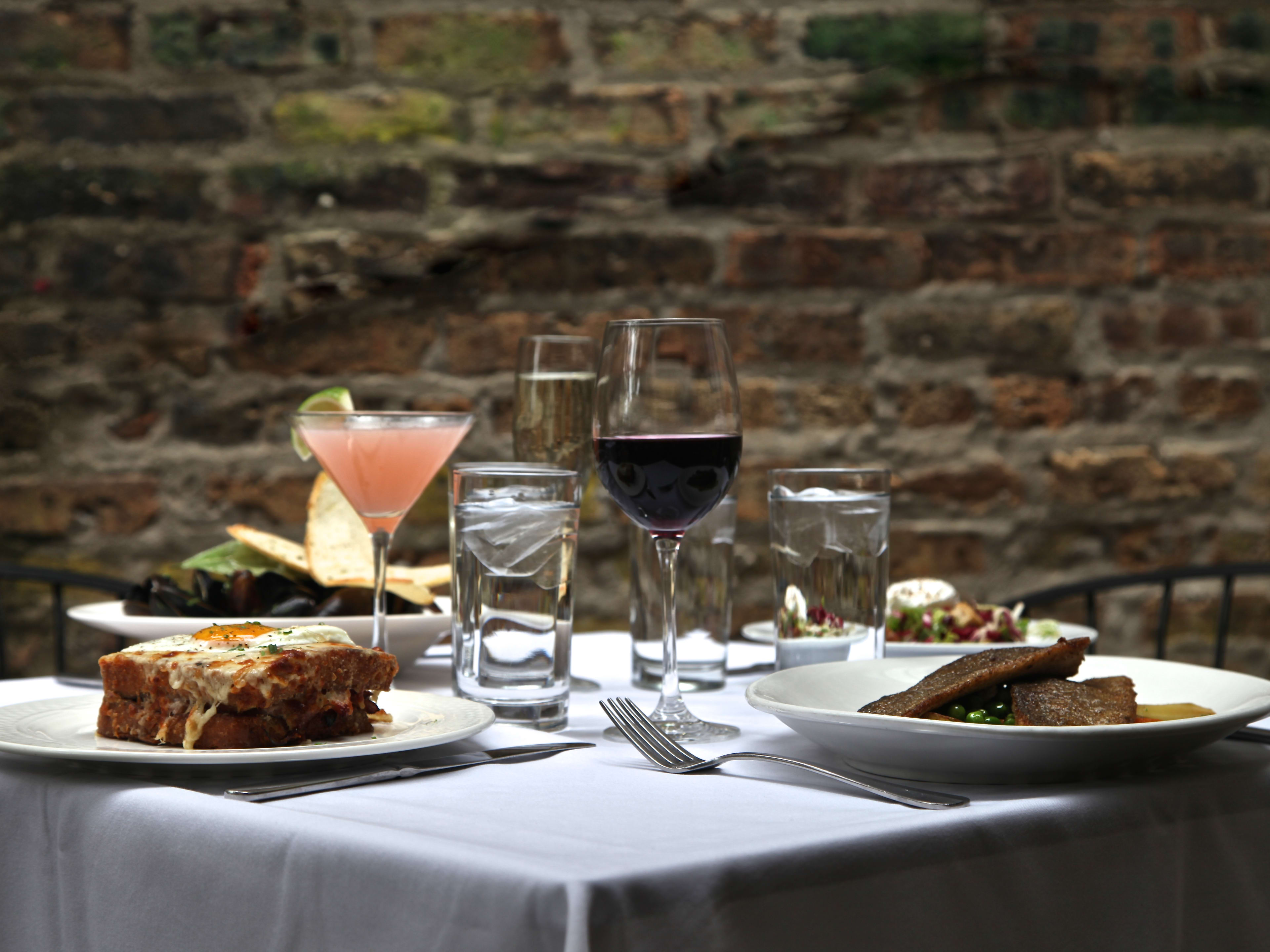 Various dishes and drinks from Bistro Campagne on a square table with a white table cloth. A brick wall is in the backgroun.