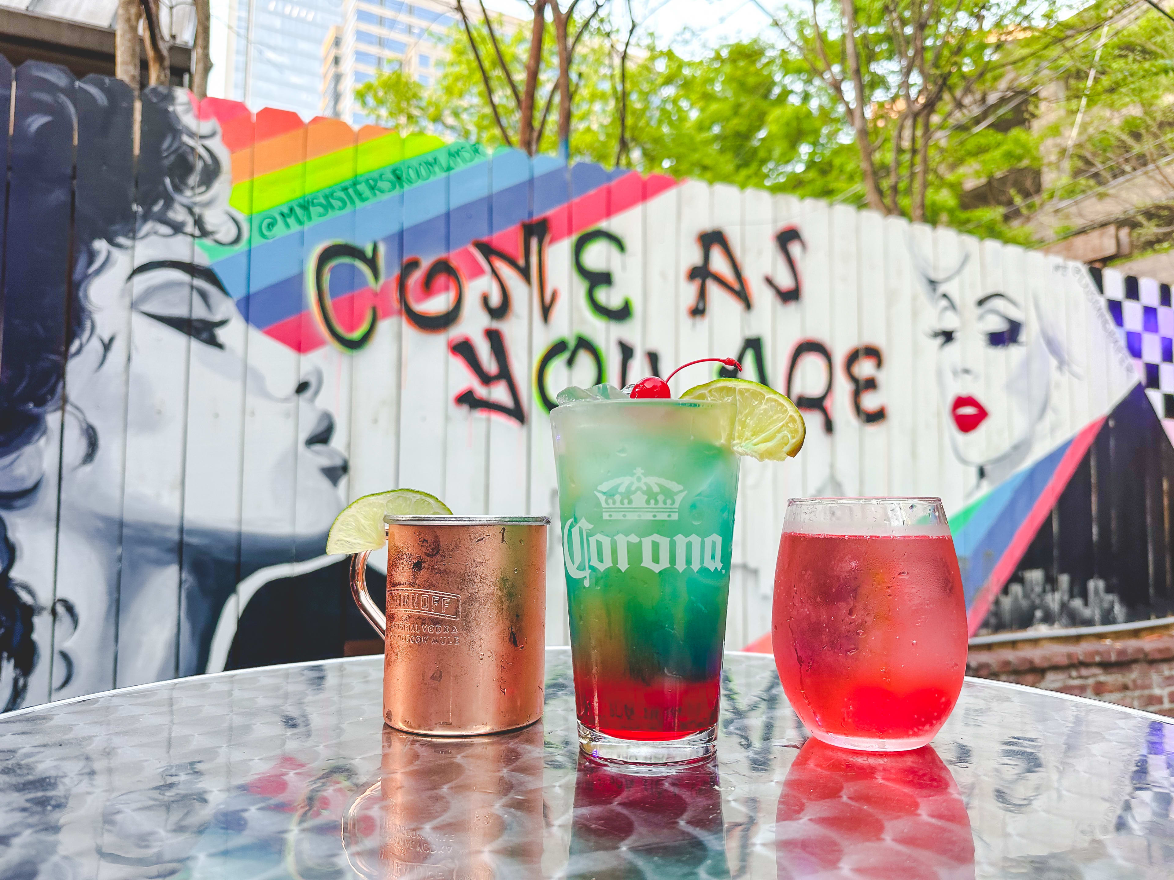 Three colorful cocktails sitting on a table in front of a mural that says "Come as you are."