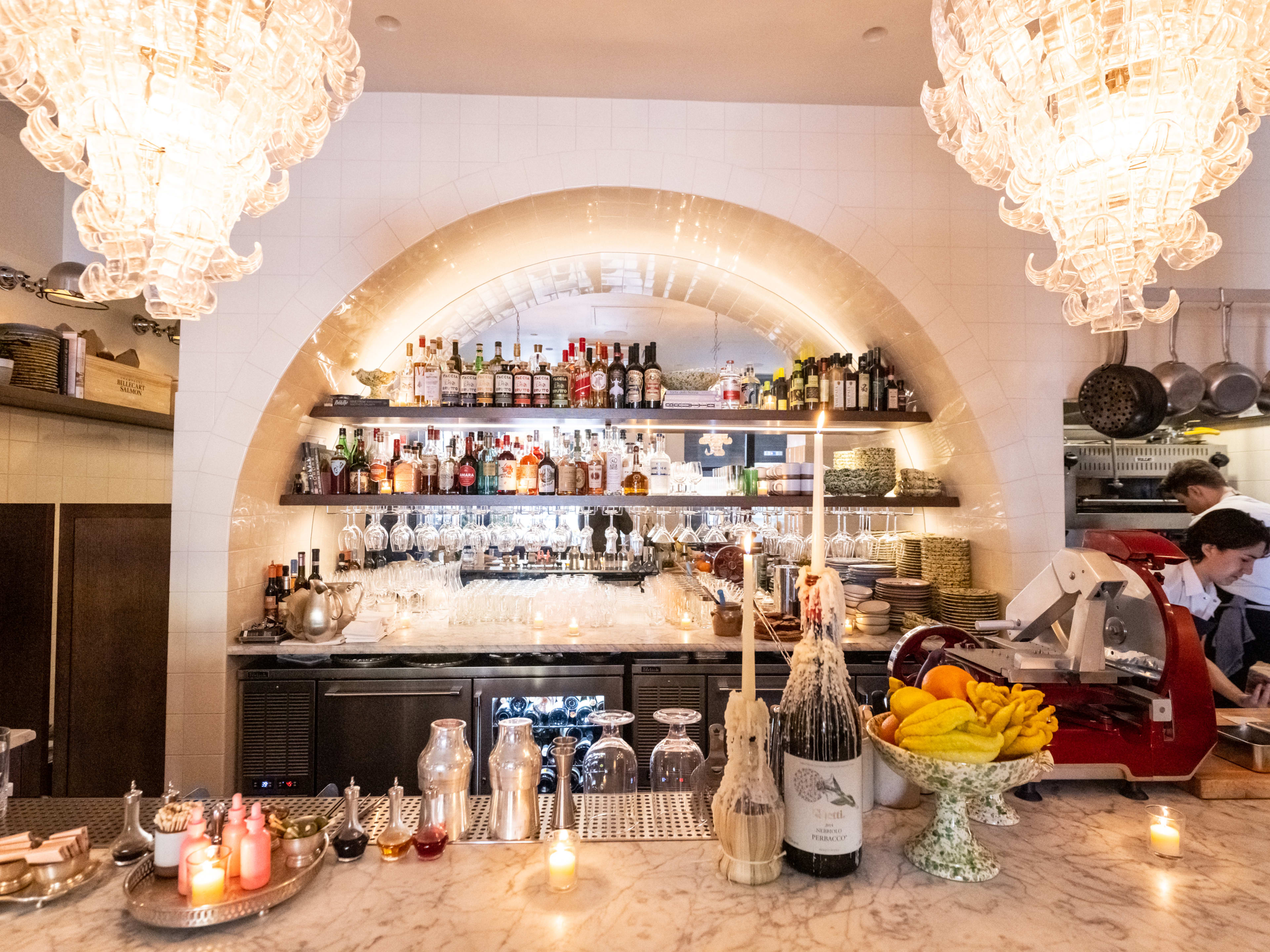 Marble bar with cured meat slicer, shelves of liquor, hanging pots and pans, and chandeliers.