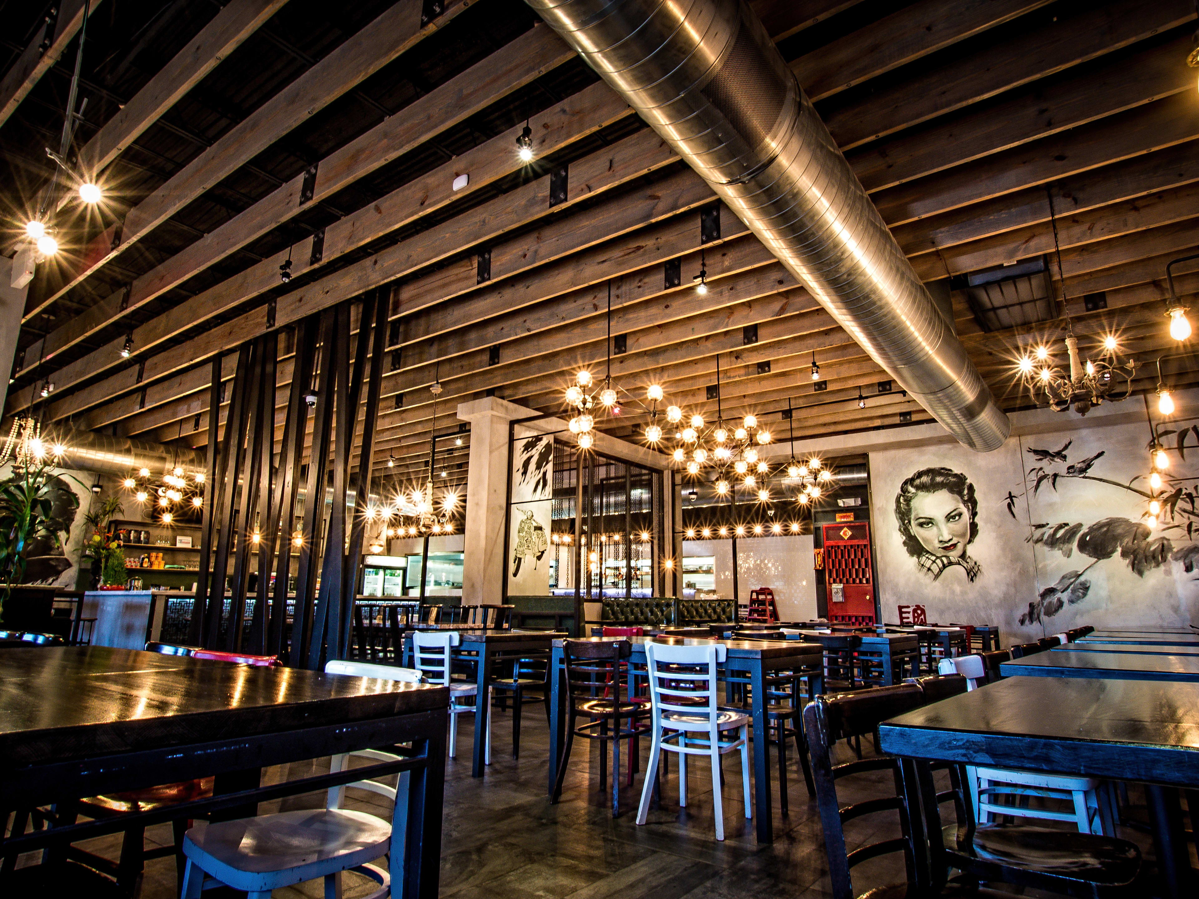 The interior of Mein. There are black and white murals of portraits painted on the walls and industrial ceiling, black rectangular high top tables and hanging pendant lights.