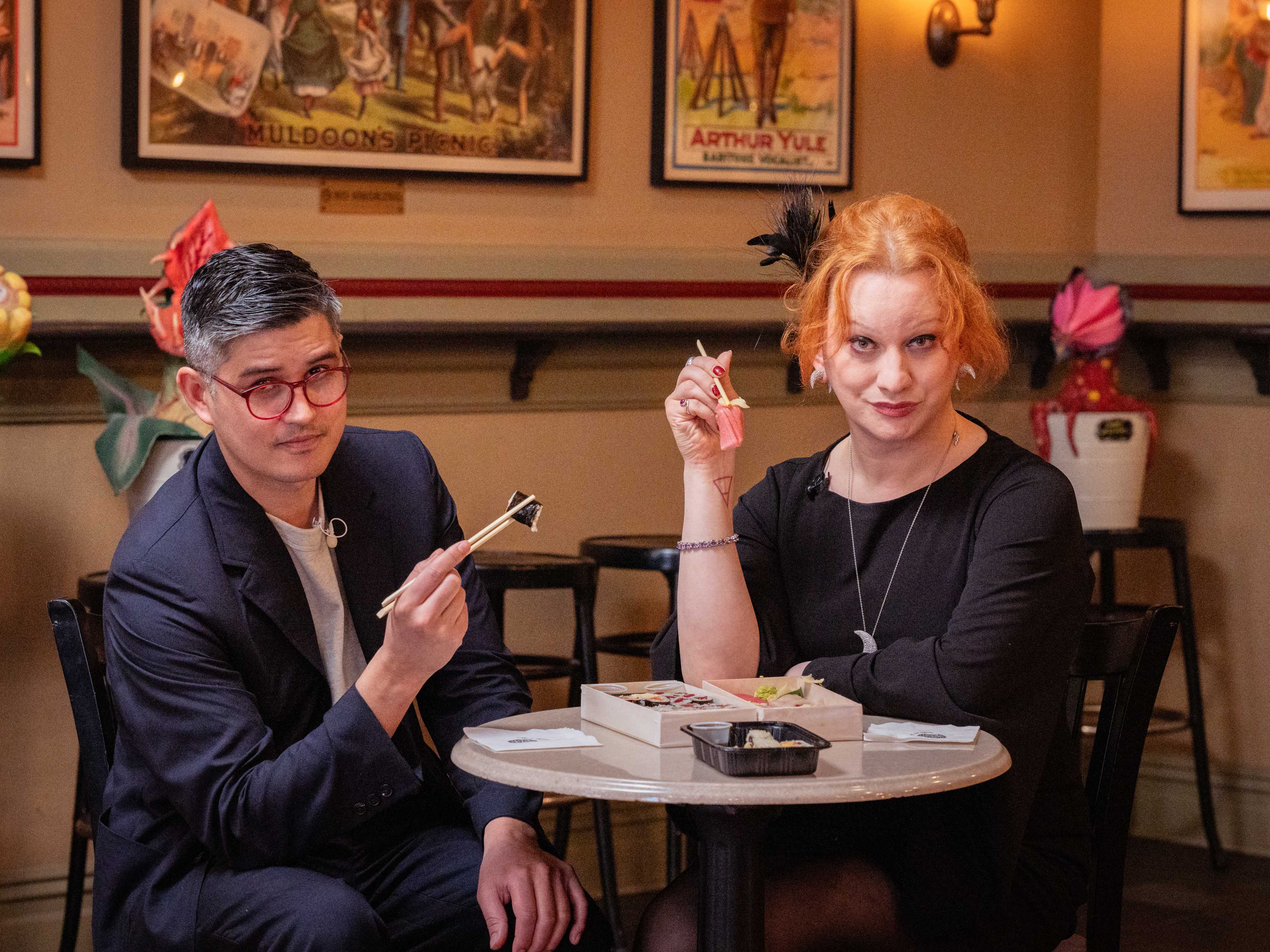 two people sit at a table. they are both holding sushi in chopsticks held out in front of them