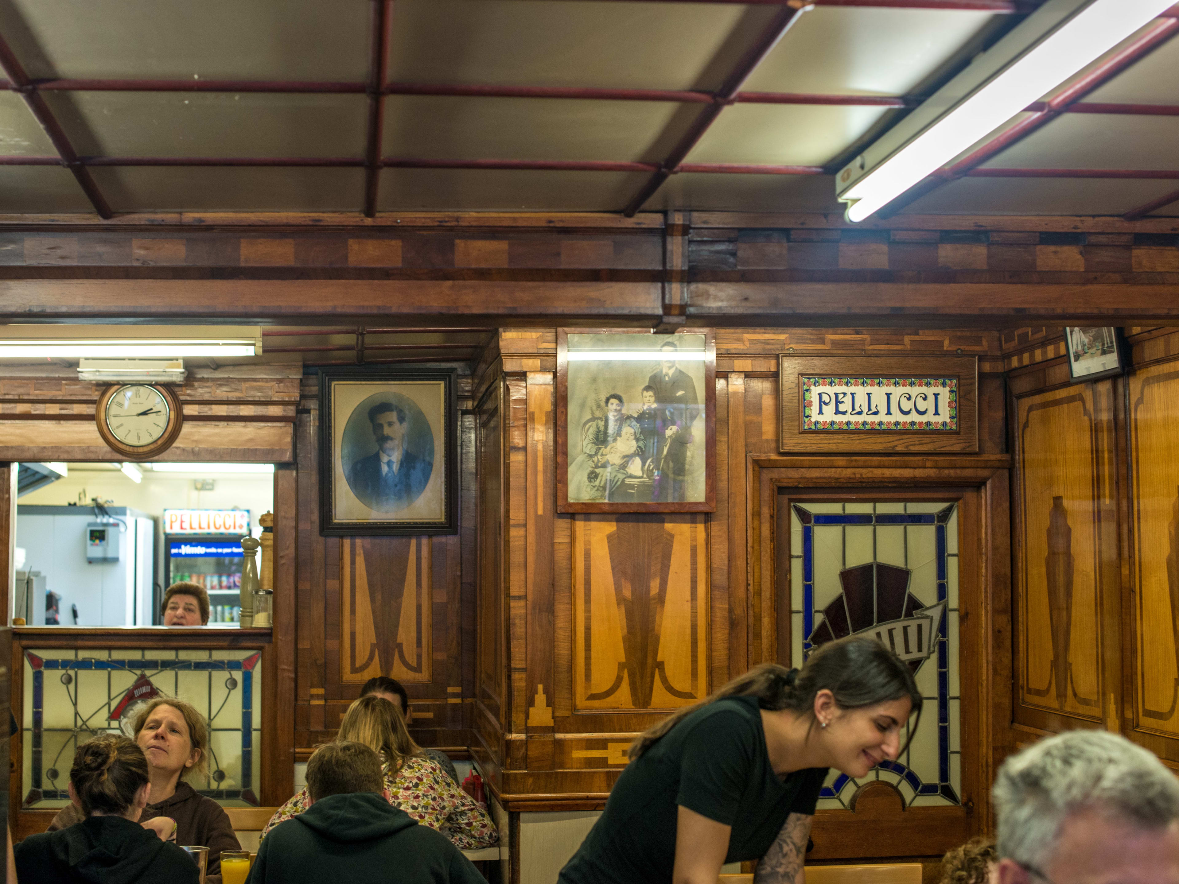 People sit at tables inside the turn of the century E. Pellicci. There are framed portraits, stained glass and detailed wooden paneling.