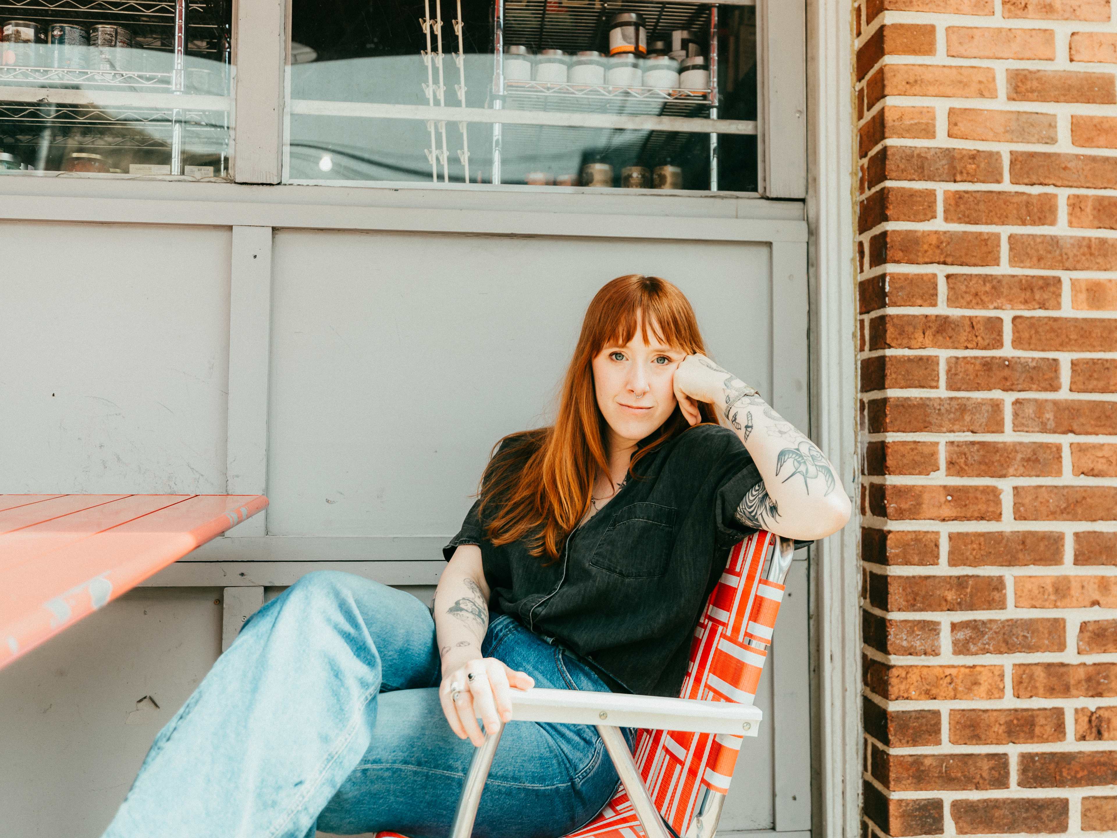 a woman with auburn hair wears a black top and blue jeans while sitting in a red chair