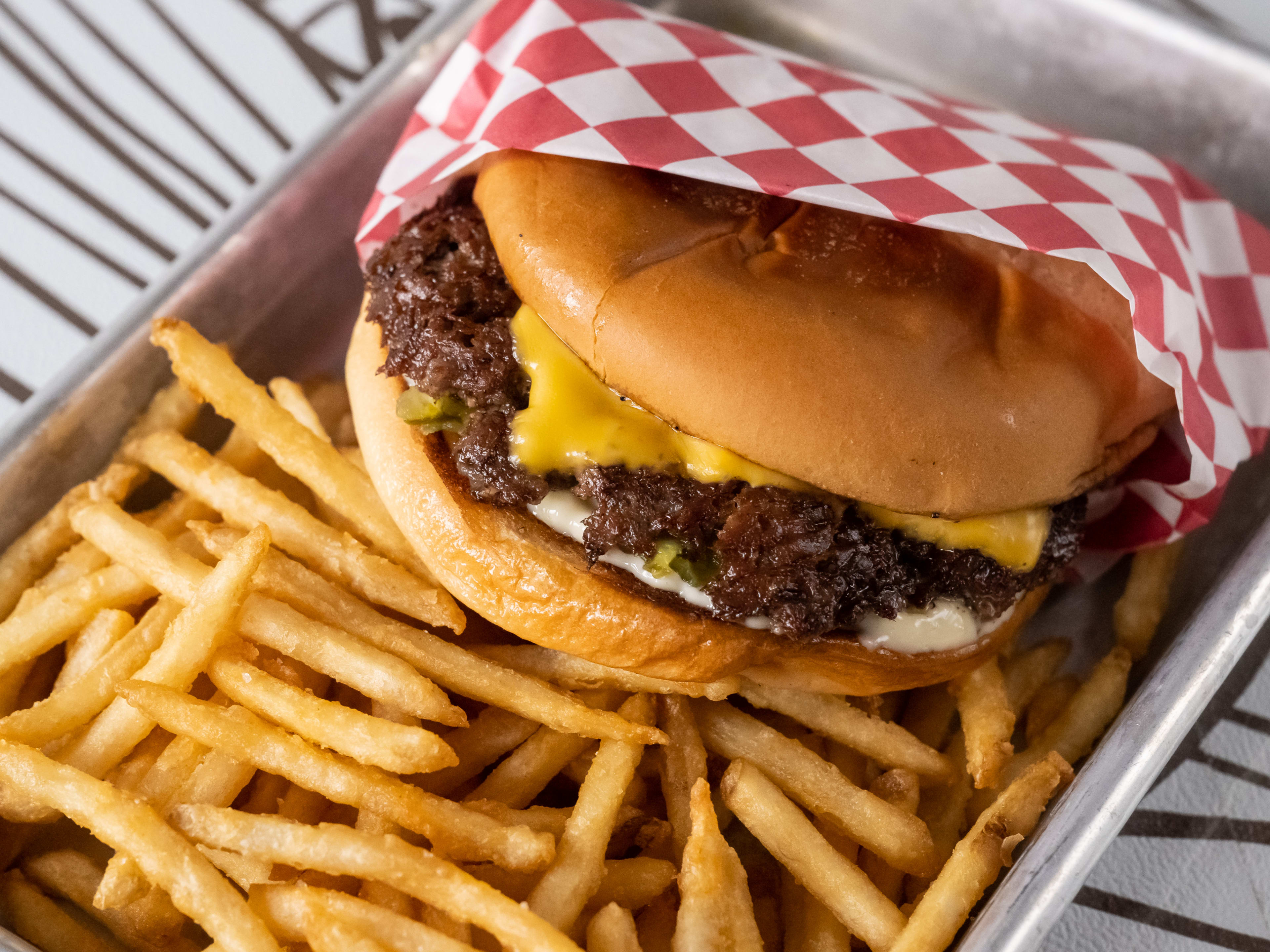 A metal tray holding a smashburger wrapped in red and white checkerboard paper on top of a pile of shoestring truffle fries.