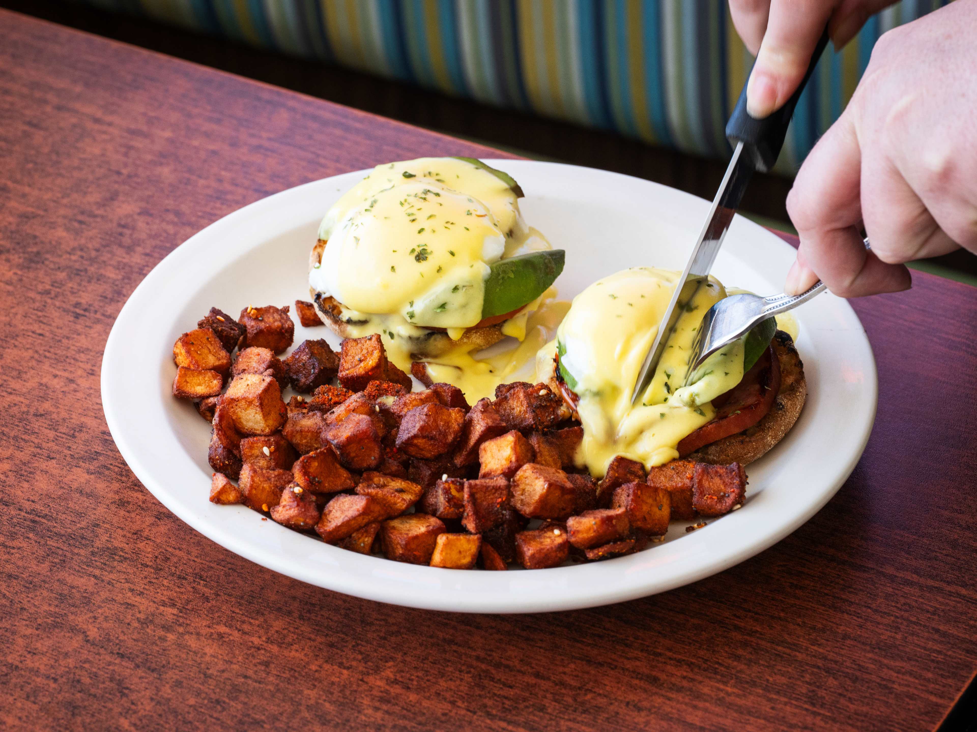 A hand cutting into a plate of eggs benedict and home fries