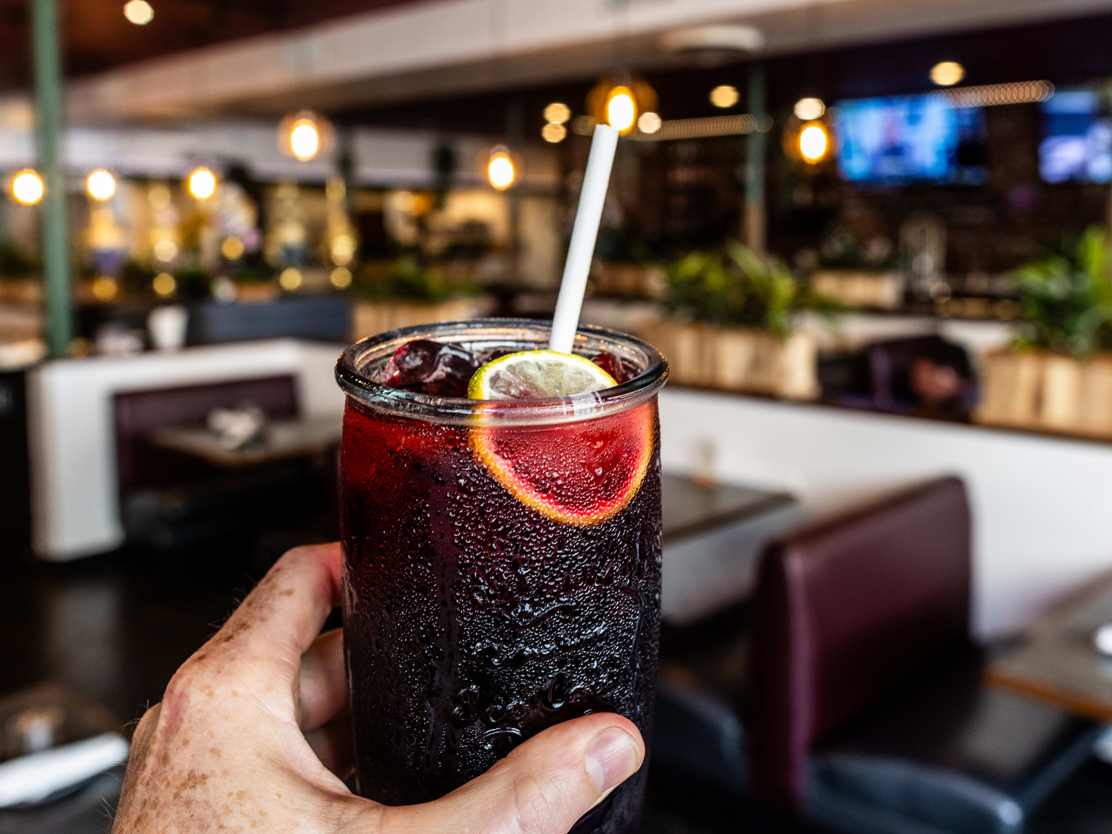 A glass of hibiscus tea with a citrus garnish held up with the interior of Khon Thai blurred in the background.