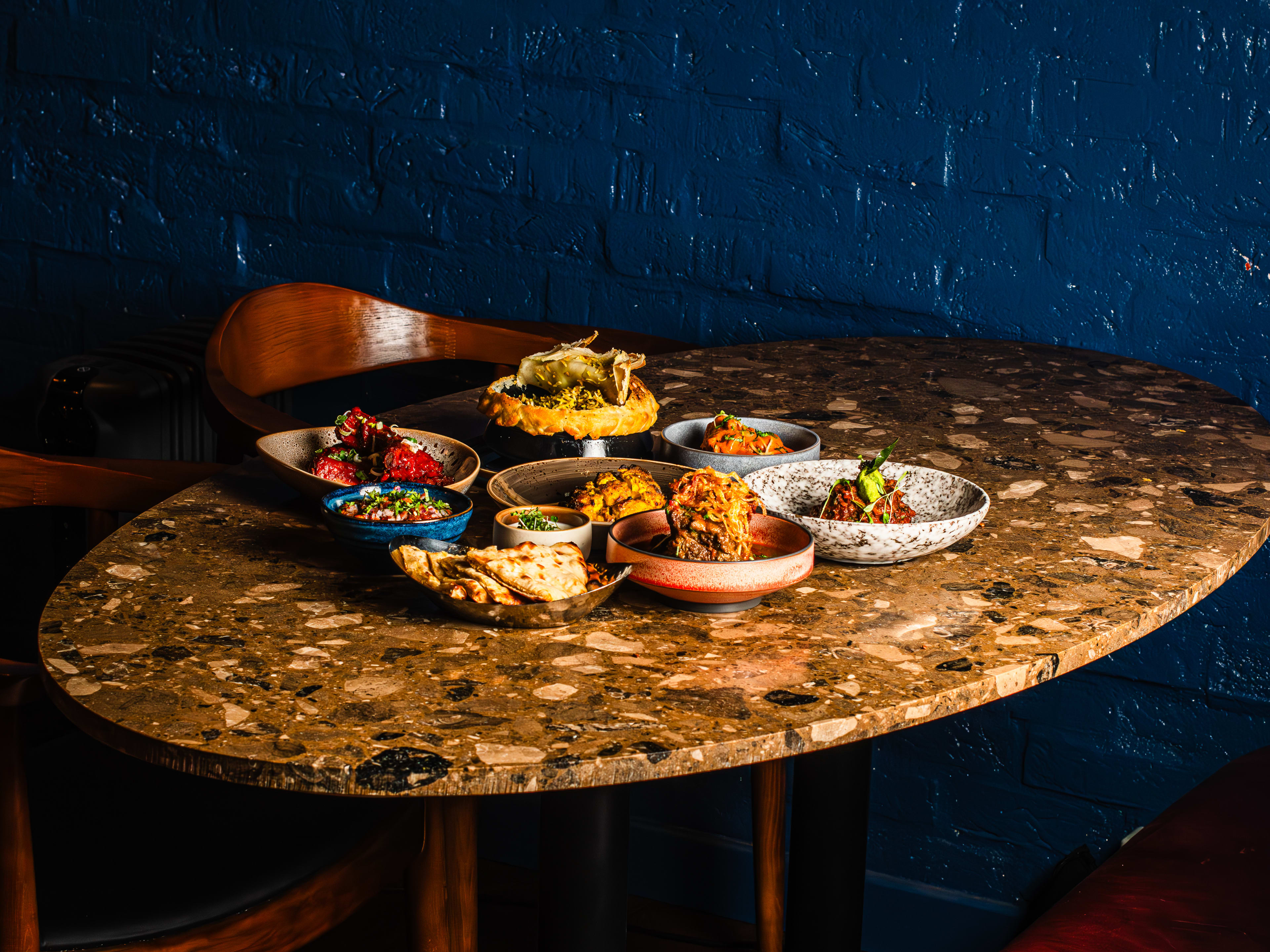 A variety of dishes from Kokum on an oval shaped granite table, set against a blue painted brick wall.