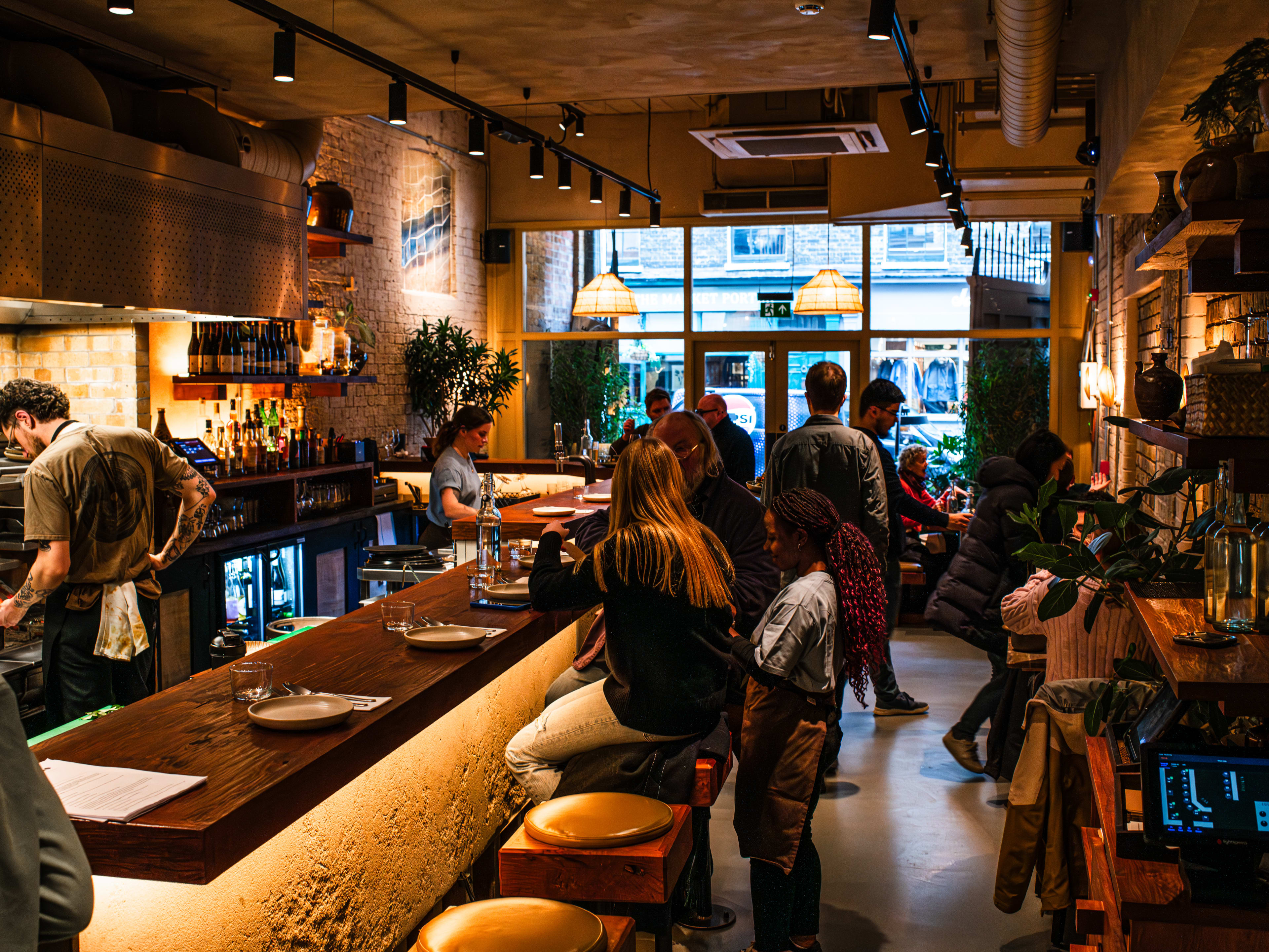 People eat at the bar and at the front tables at Kolae. A chef is seen cooking on the grills behind the bar.