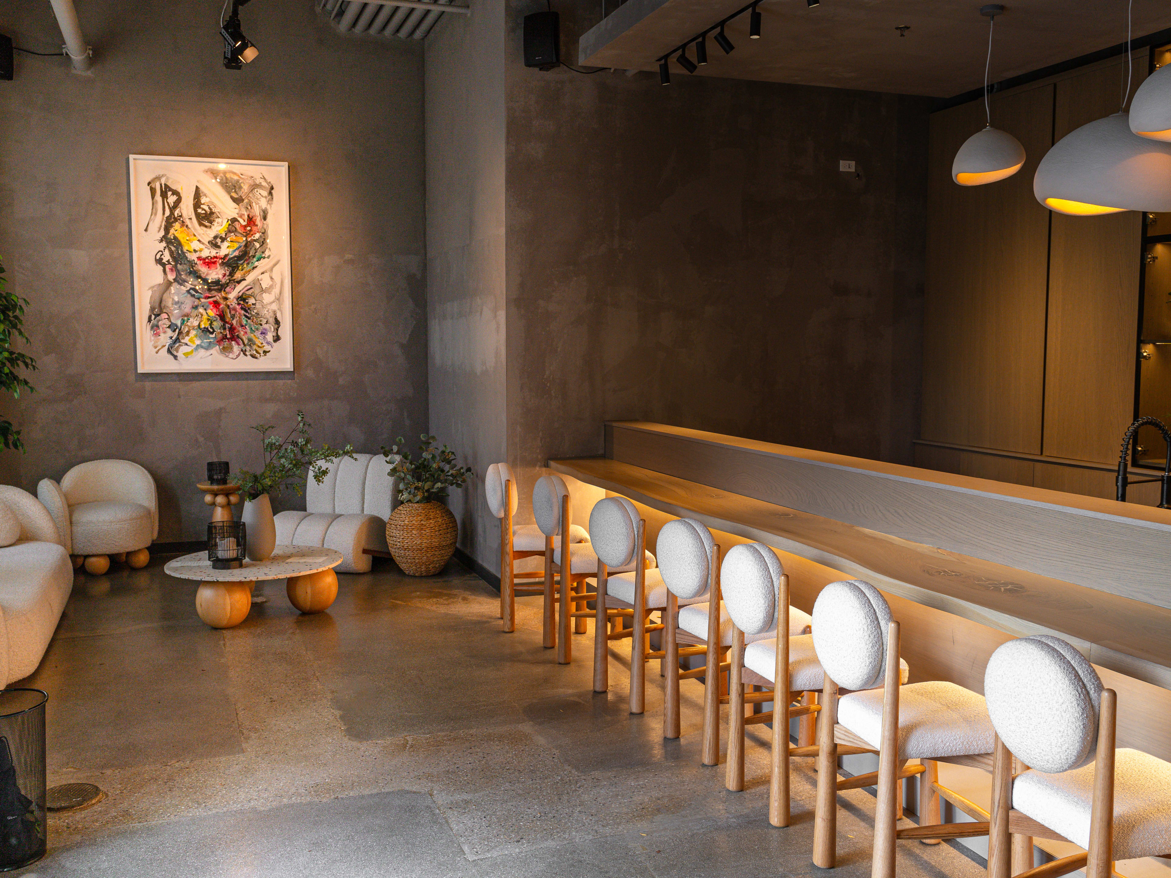 The grey slate interior of Kyoten Next Door. The sushi bar is to the right, lined with white chairs.  There are white ovular hanging pendants. There is a seating area in the back corner with plants, white mod furniture and a low white coffee table.