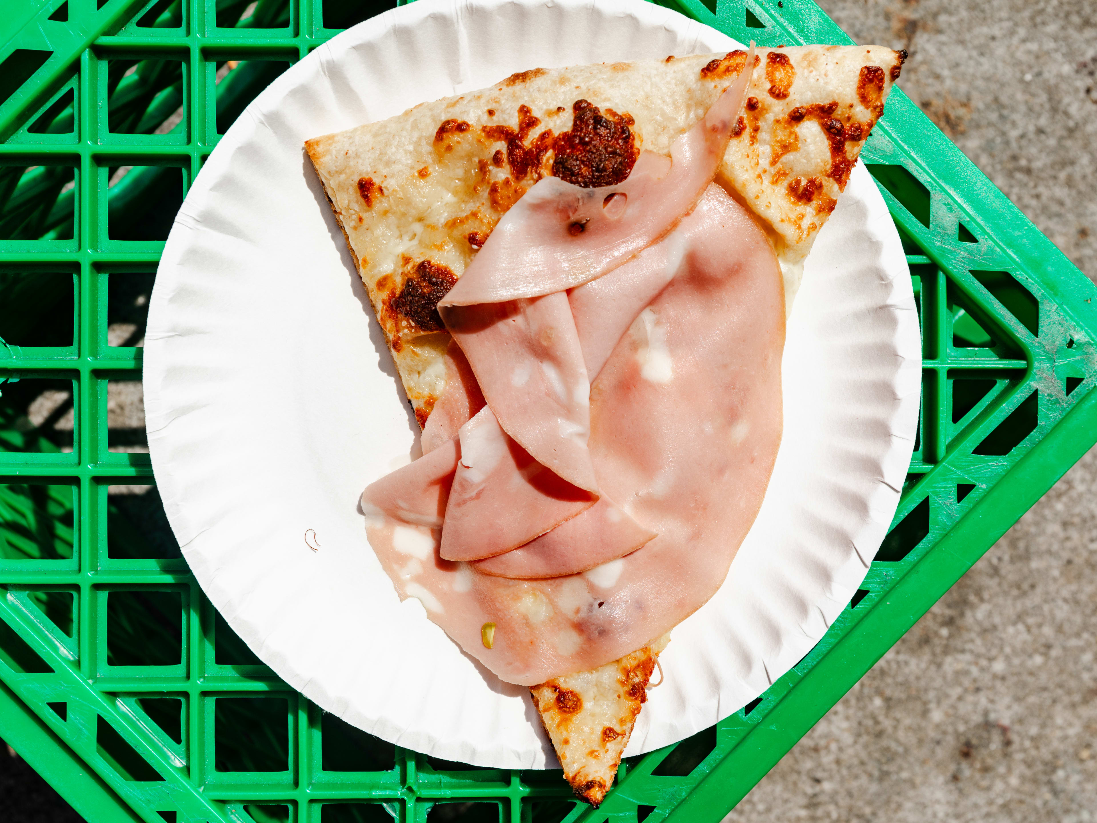 A slice of mortadella pizza on a paper plate on top of a green milk crate.