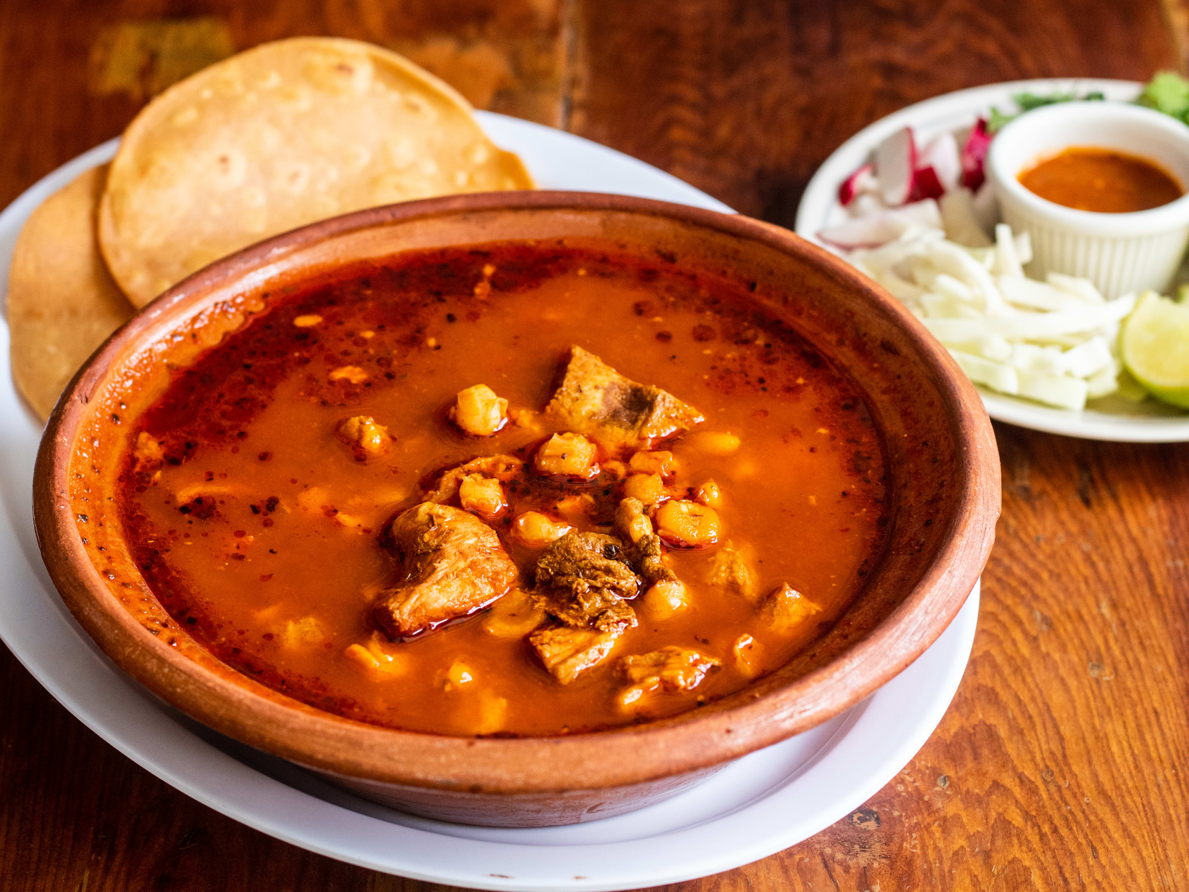 Bowl of pozole with tortillas and a side dish of limes and sauce.