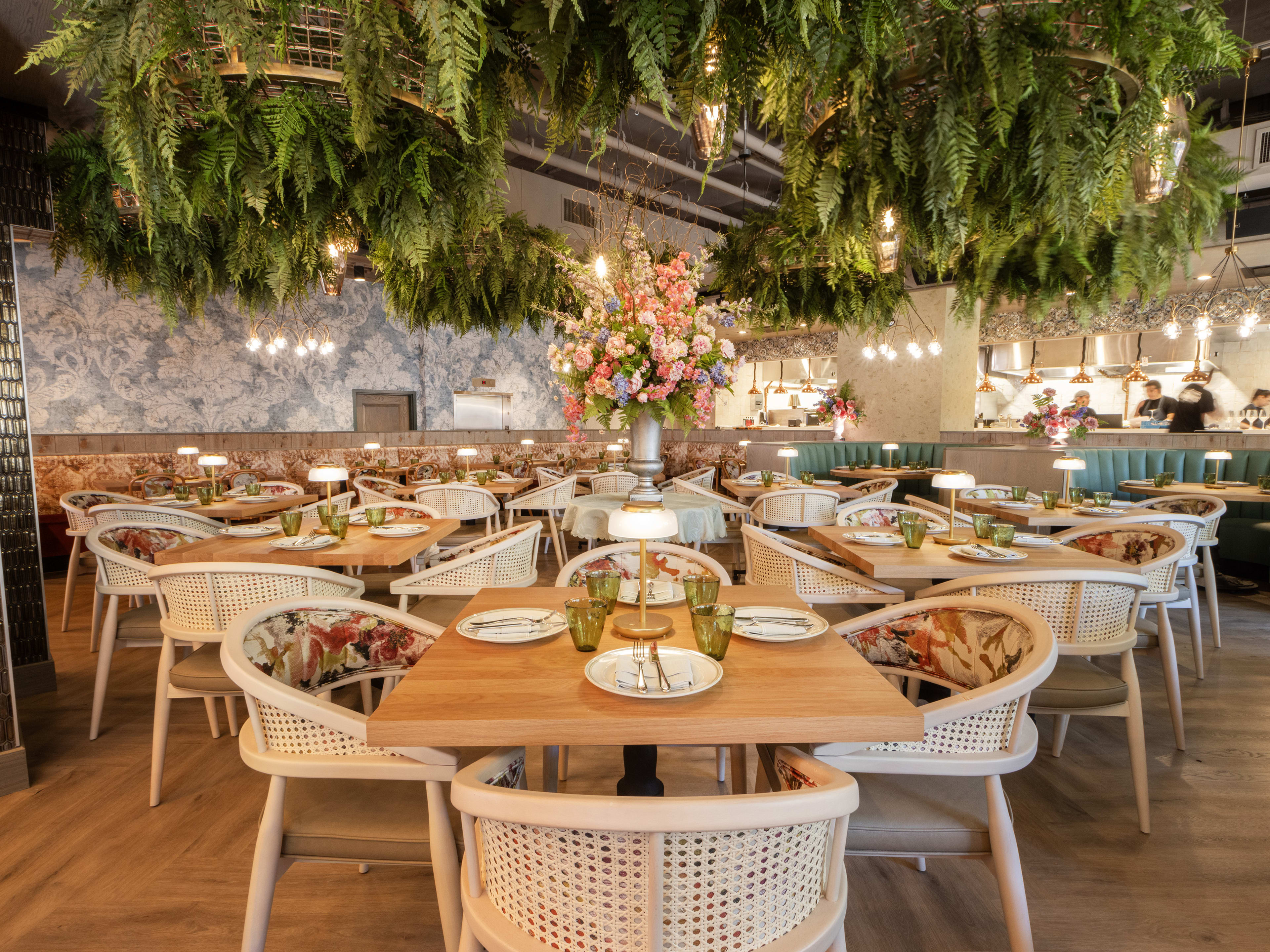 A bright dining room full of tables with plants hanging from the ceiling and an open kitchen in the background.