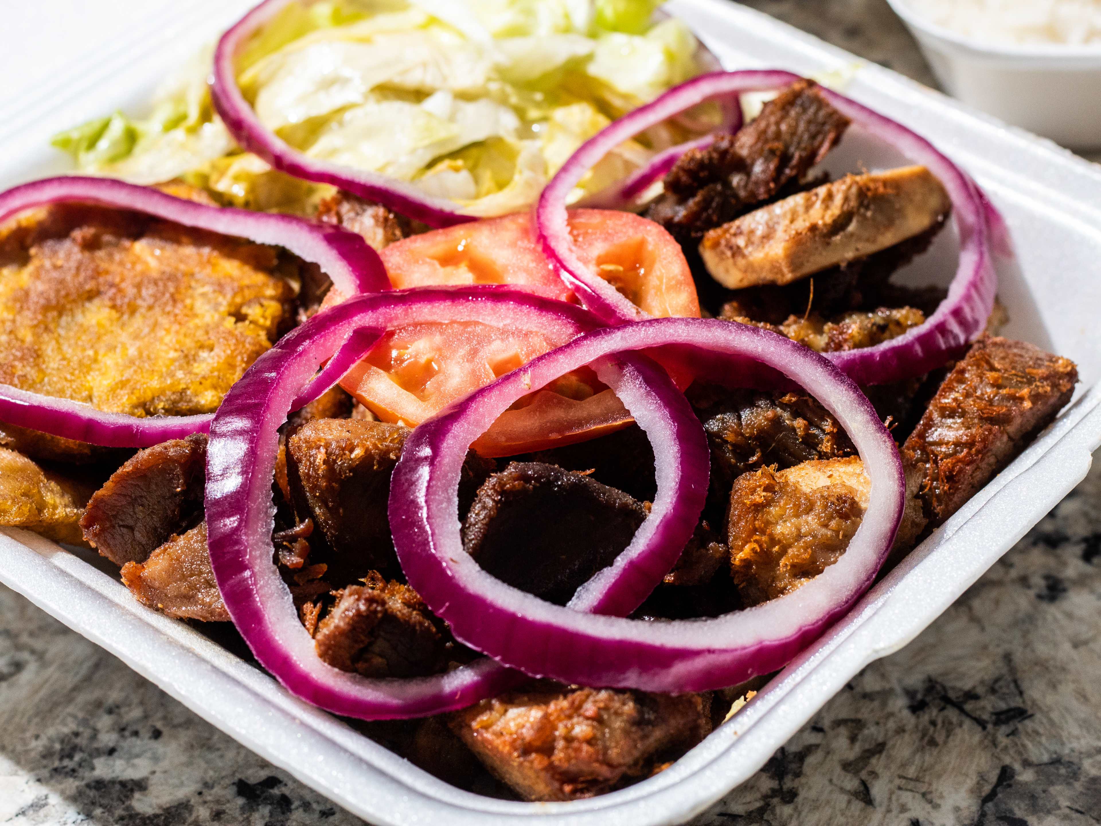 A takeout box of griot with rings of red onion on top.