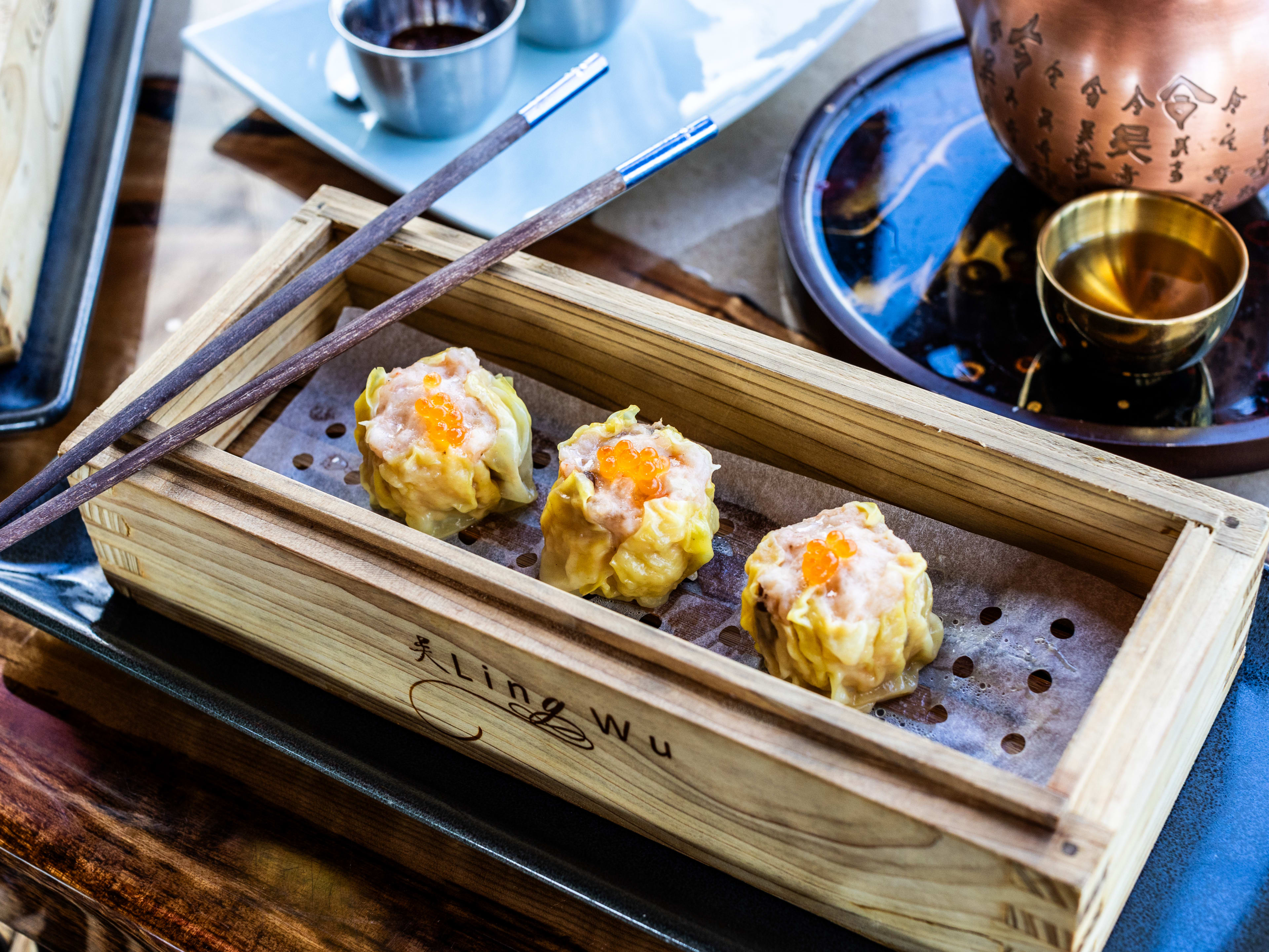 A wooden steamer box with three scallop and caviar siu mai. A copper tea pot is in the background.