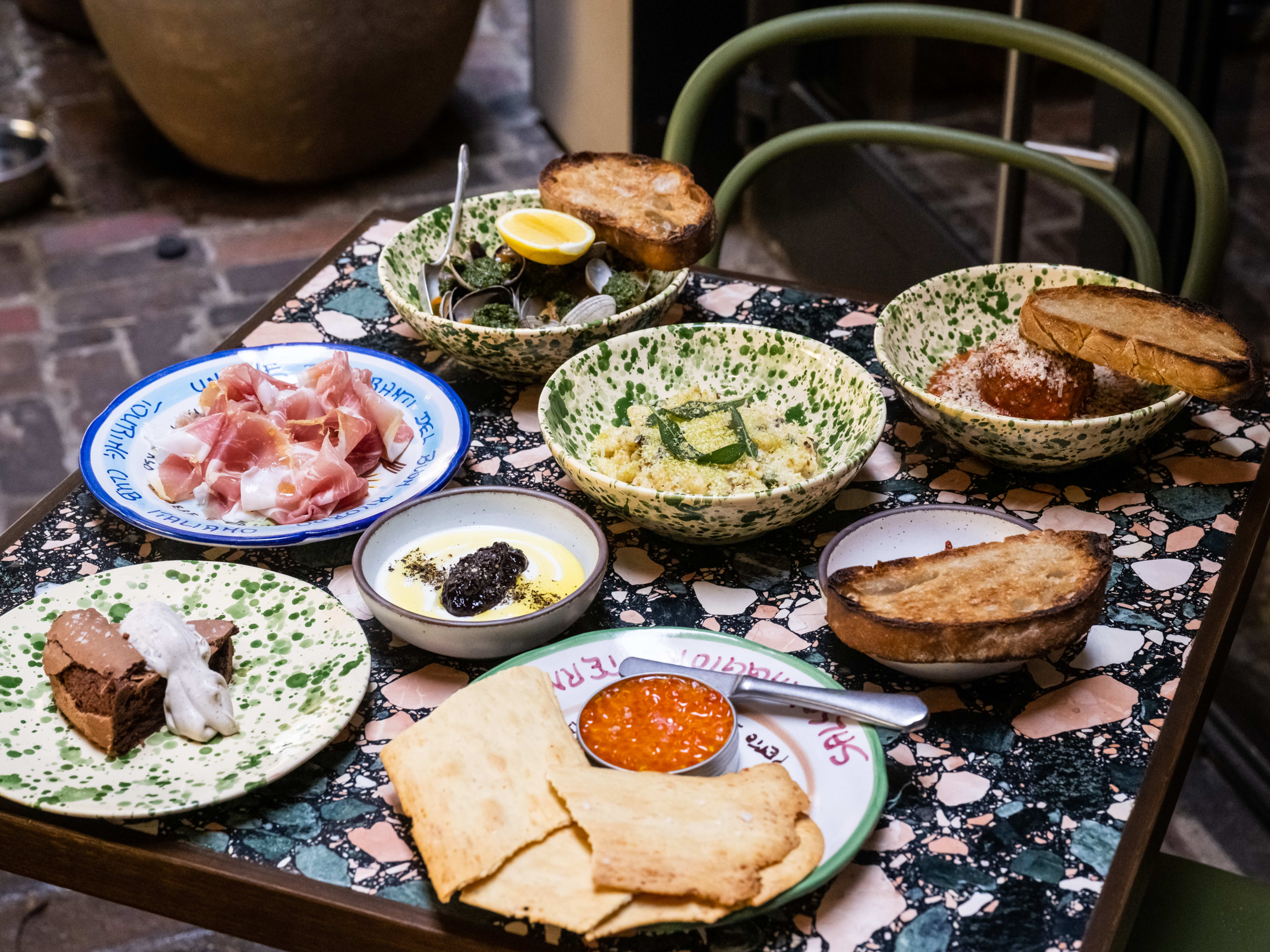 A spread of Italian snacks, like whipped cod topped with ikura, cavatelli, a meatball, a bowl of clams, prosciutto, panna cotta, and a dark chocolate cake topped with mascarpone cream.