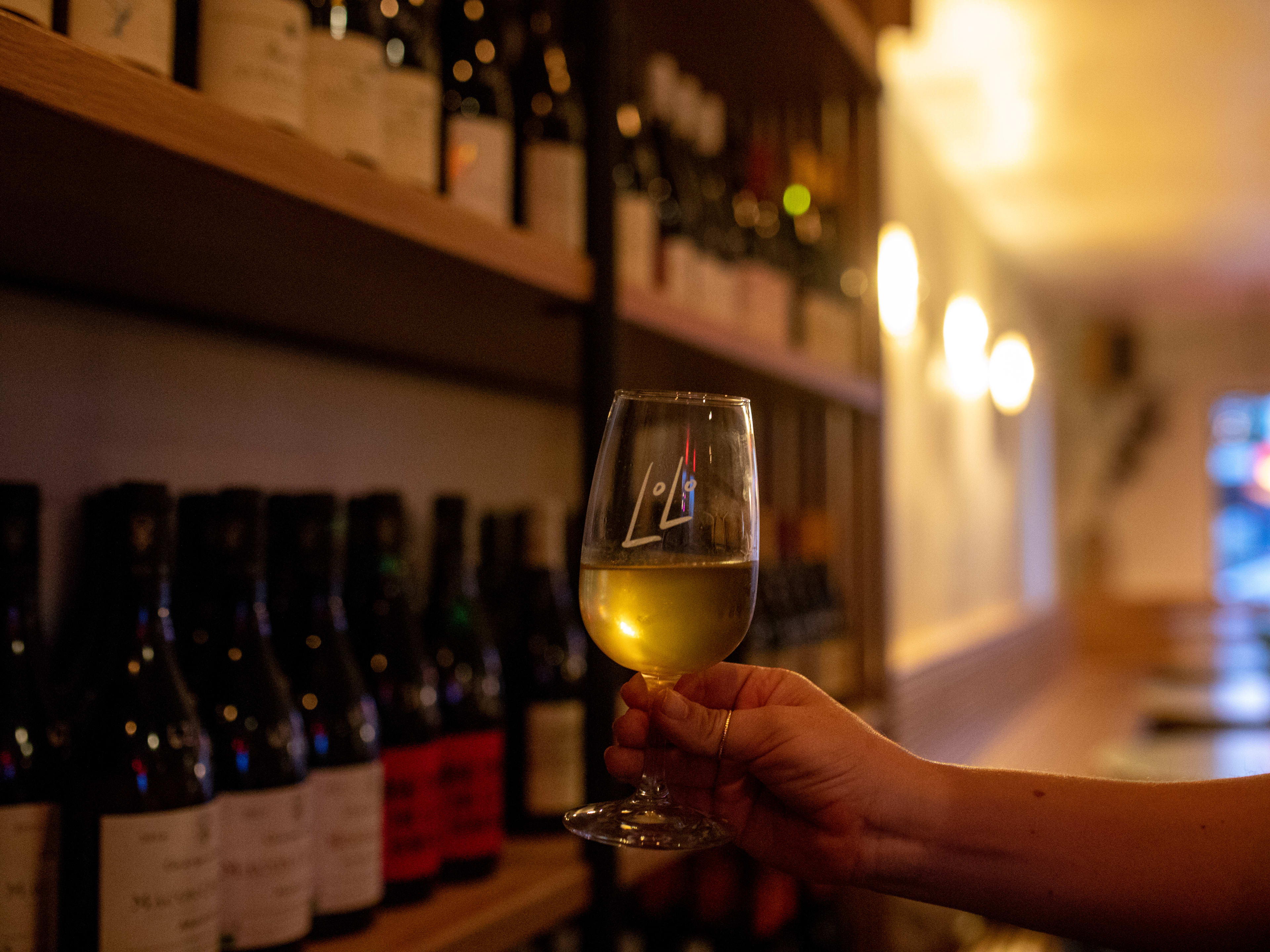 A person holds a branded glass of white wine near the shelves of bottles for purchase at LoLo.