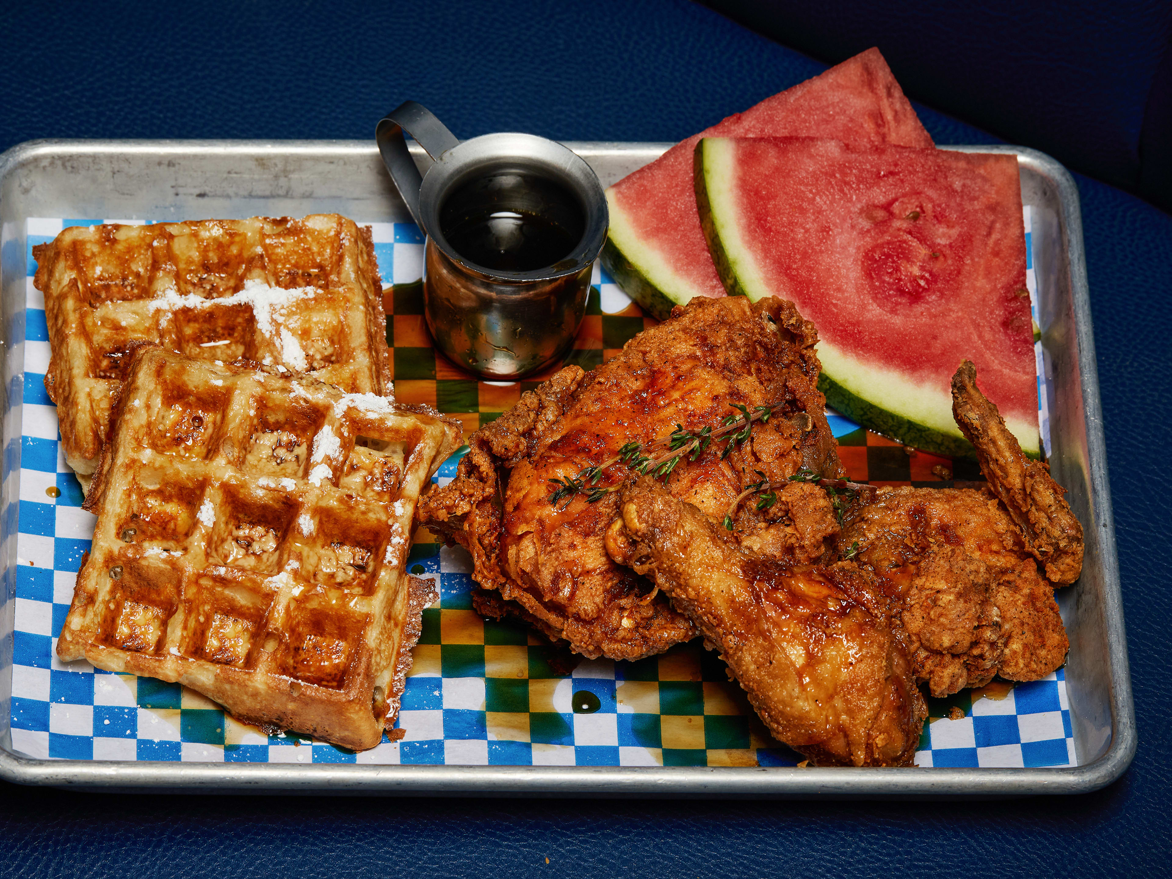 Square waffles, fried chicken, watermelon triangles, and a little tin pitcher of syrup on a checkered paper lined metal tray.