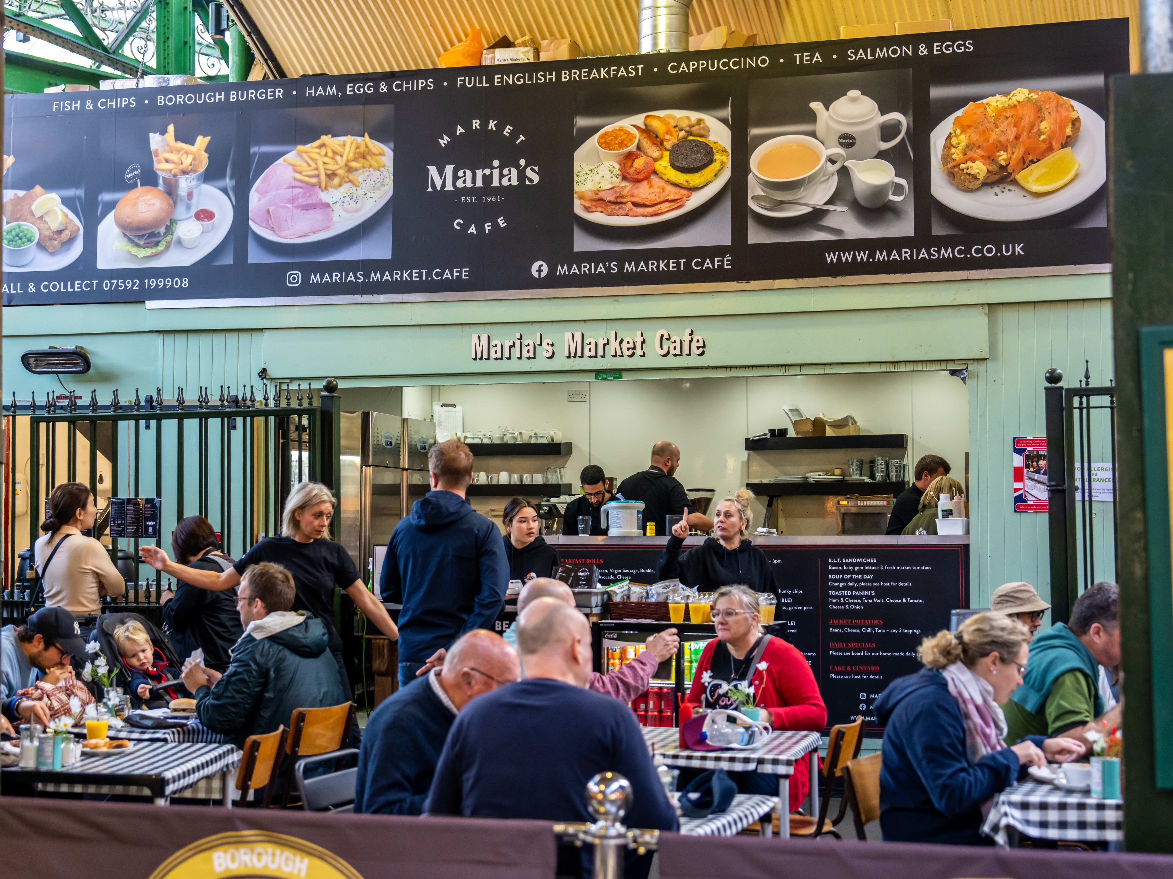 People dining at tables with checkered tablecloths and people stand at the ordering counter for Maria's Market Cafe. A banner with photos of the breakfast offerings hangs above the order counter.