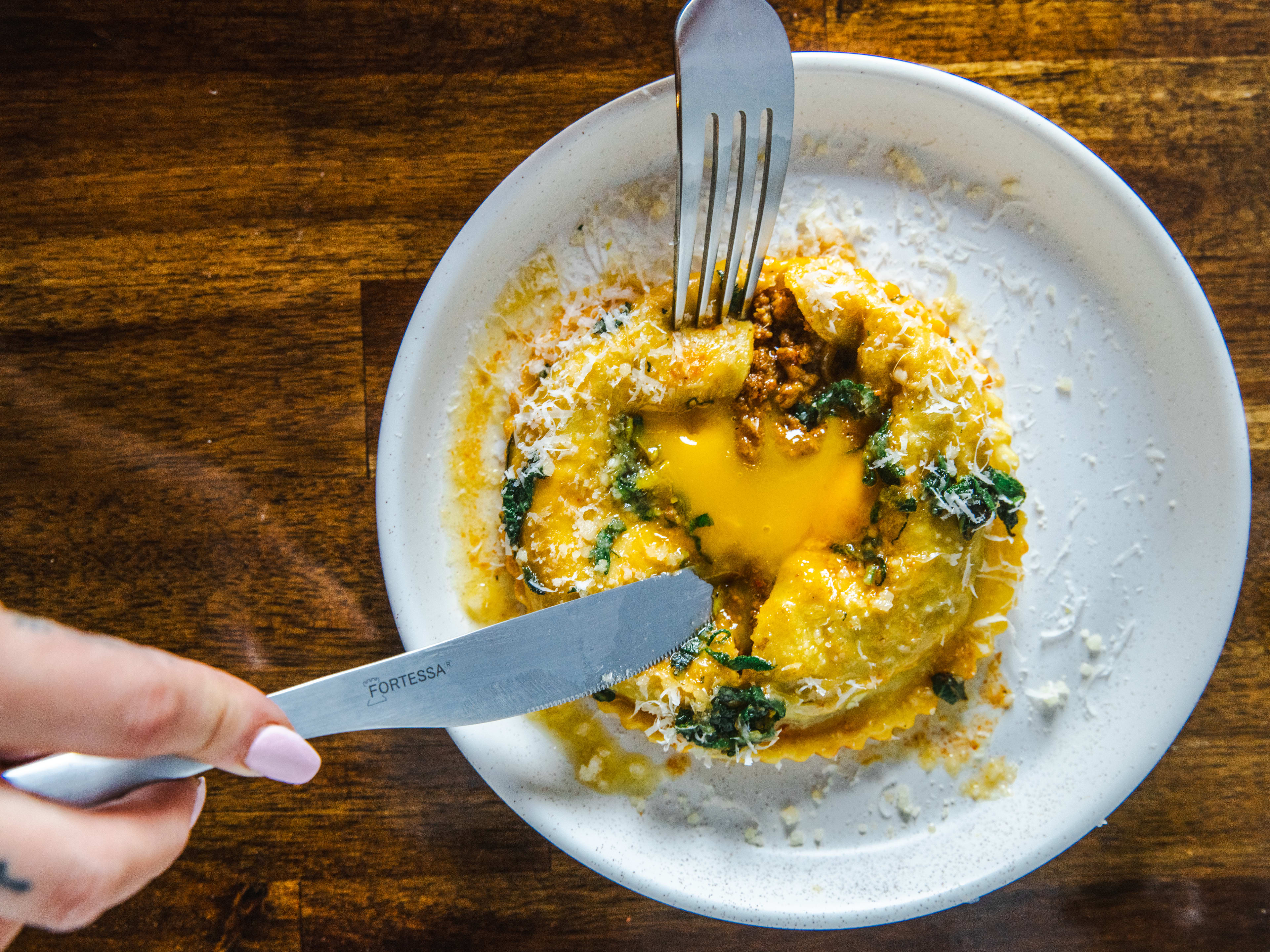 Two hands with cutting into a chopped cheese raviolo with a fork and knife.