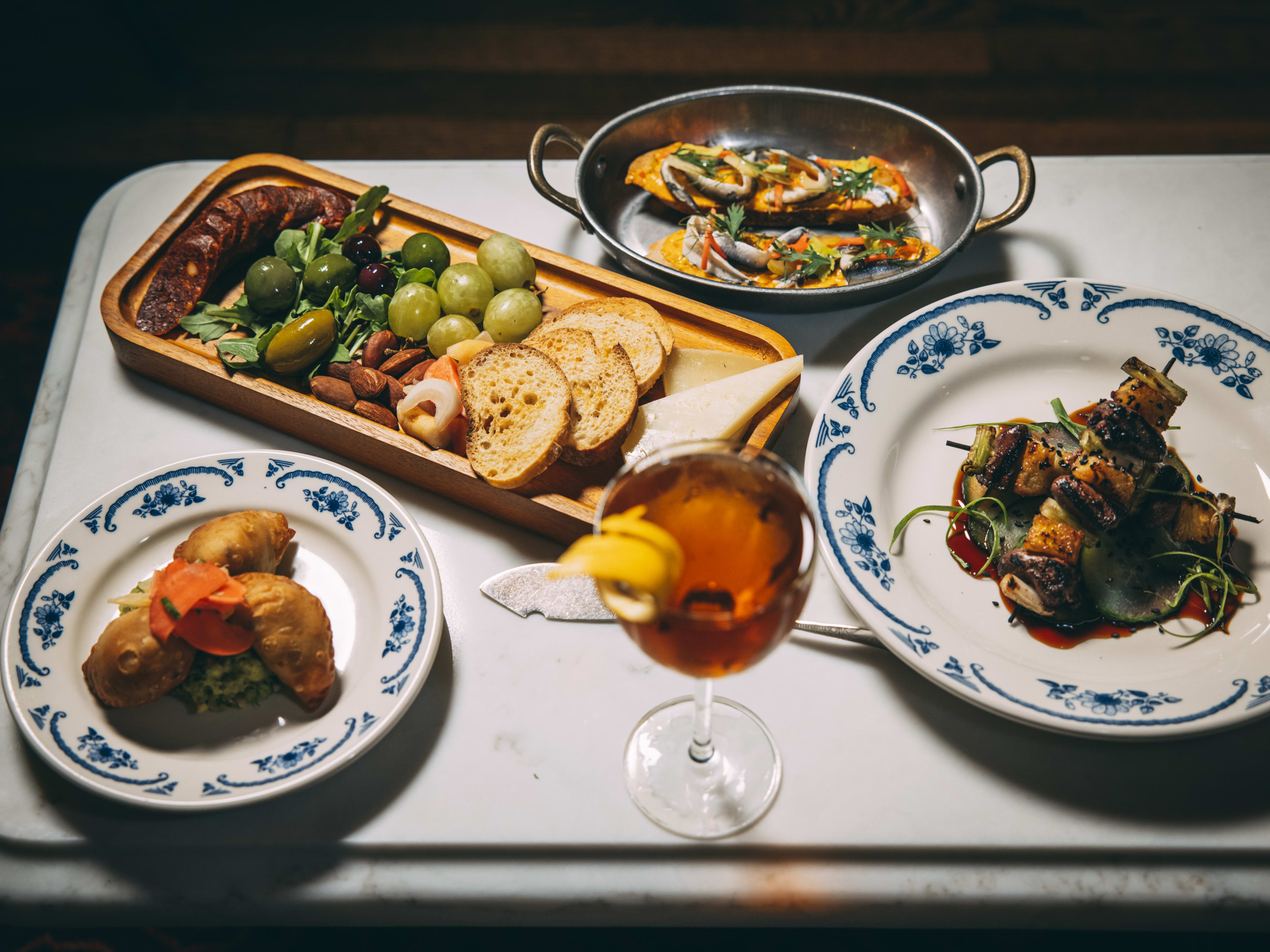 A food spread with mini empanadas, grilled chicken hearts, bread, olives, and a cocktail.