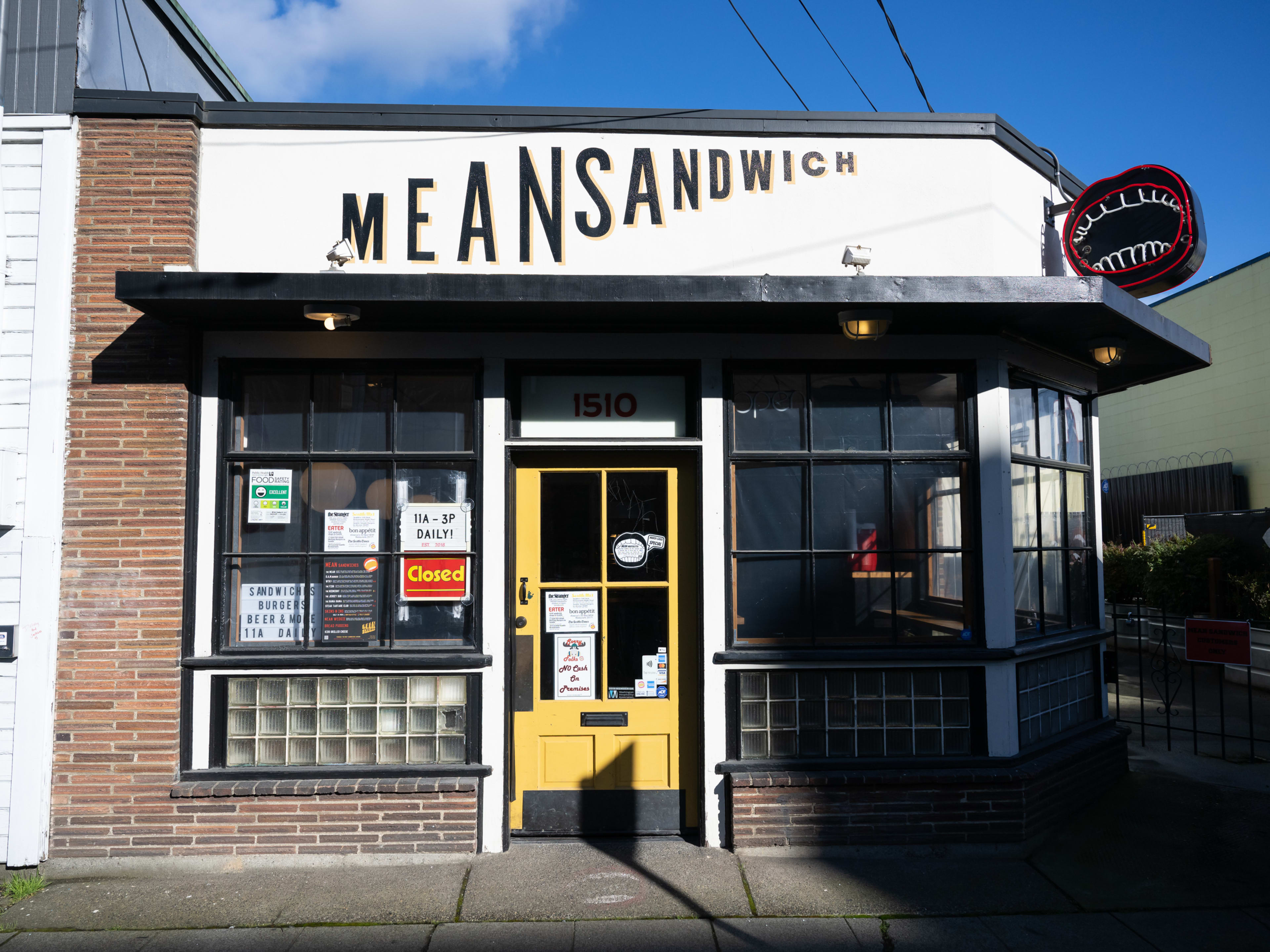 Exterior of Mean Sandwich, with white siding, exposed brick, and a neon sign depicting an open mouth.