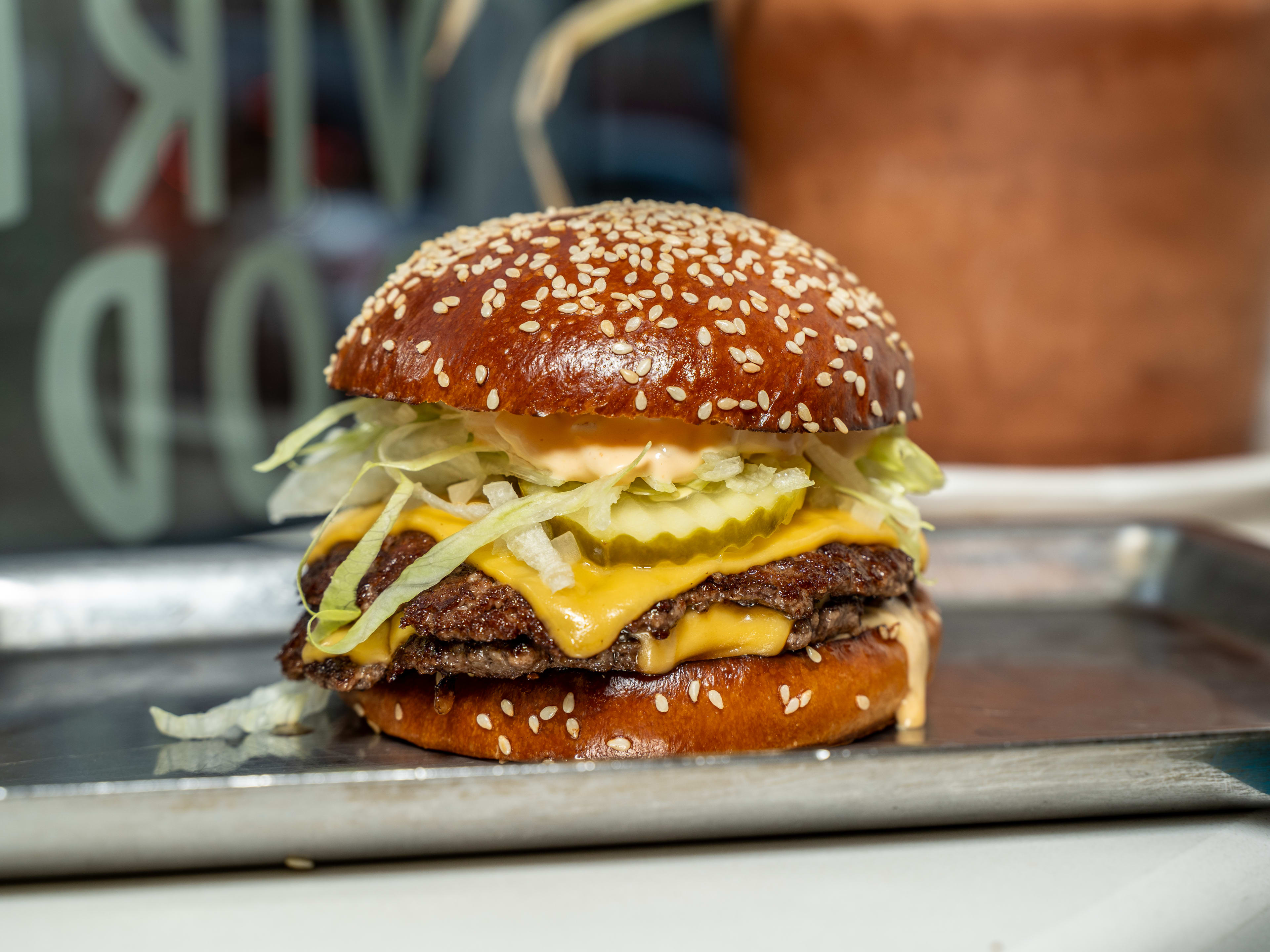 burger with seeded bun, crinkle cut pickles, shredded lettuce, two patties, and melty cheese on a metal tray