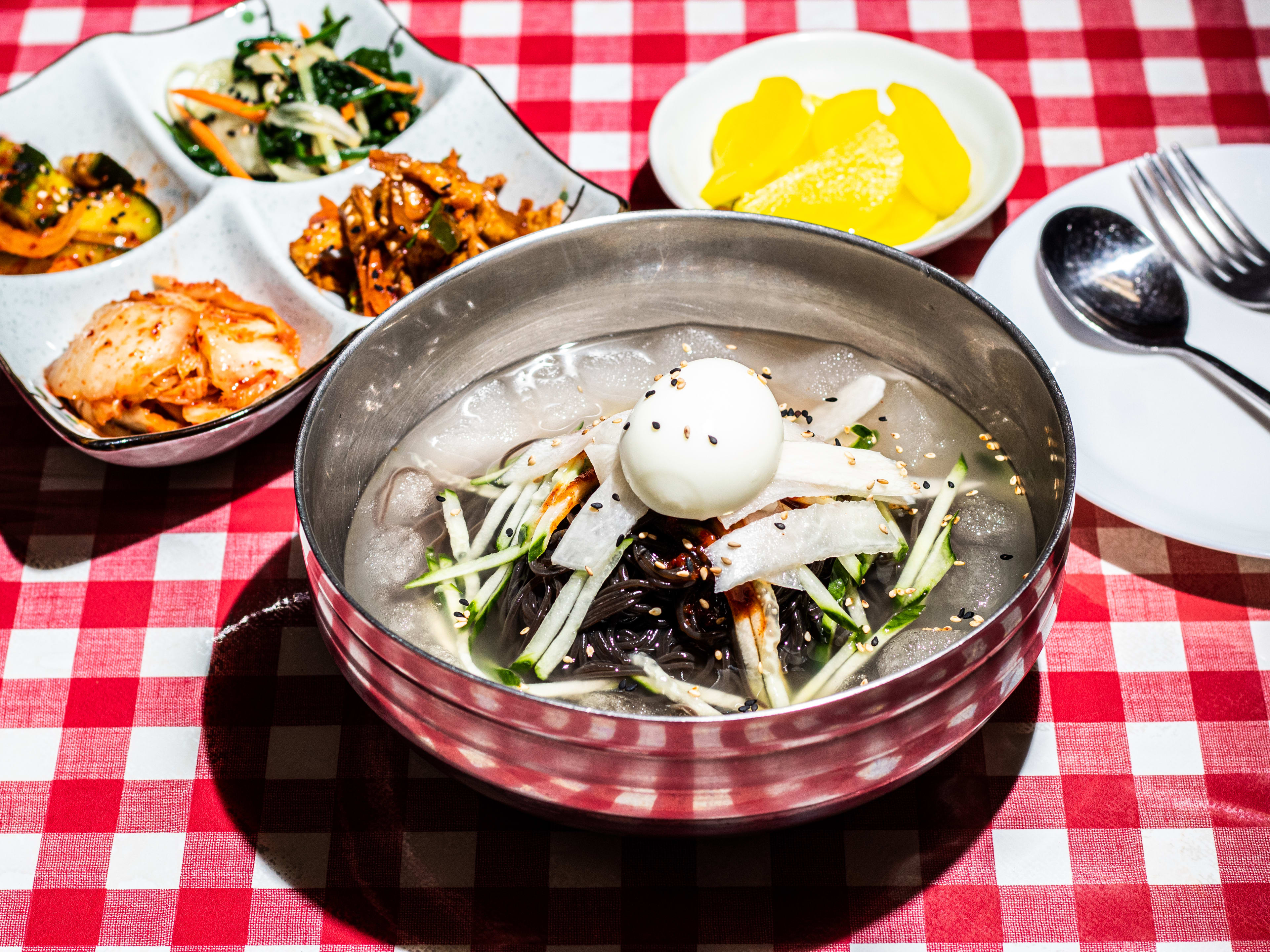 Spread of dishes including kimchi/banchan, cold noodle dish in tin bowl with whole egg, and cutlery.