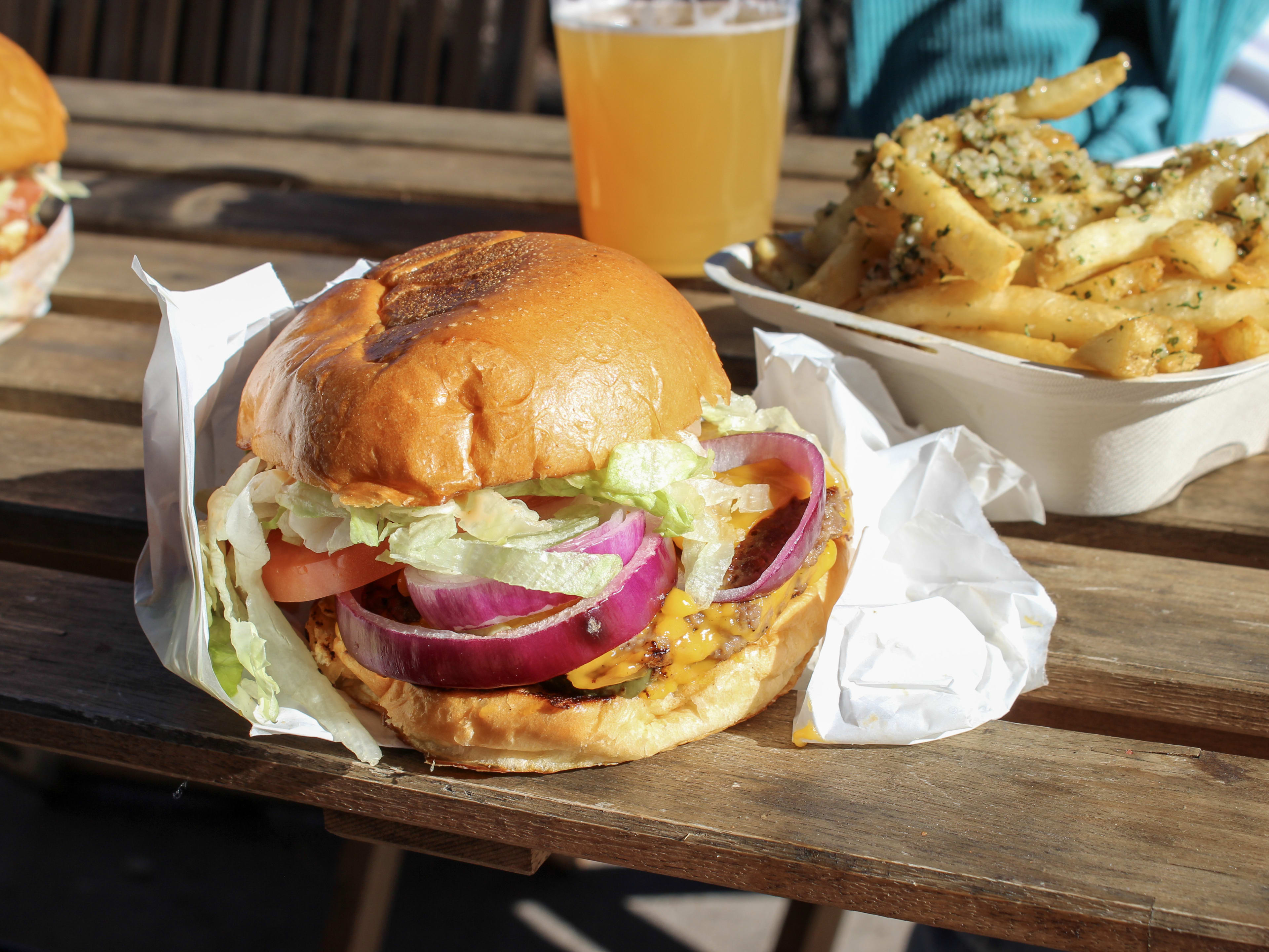 A burger wrapped in paper sits on a wooden table with fries and a beer in the background.