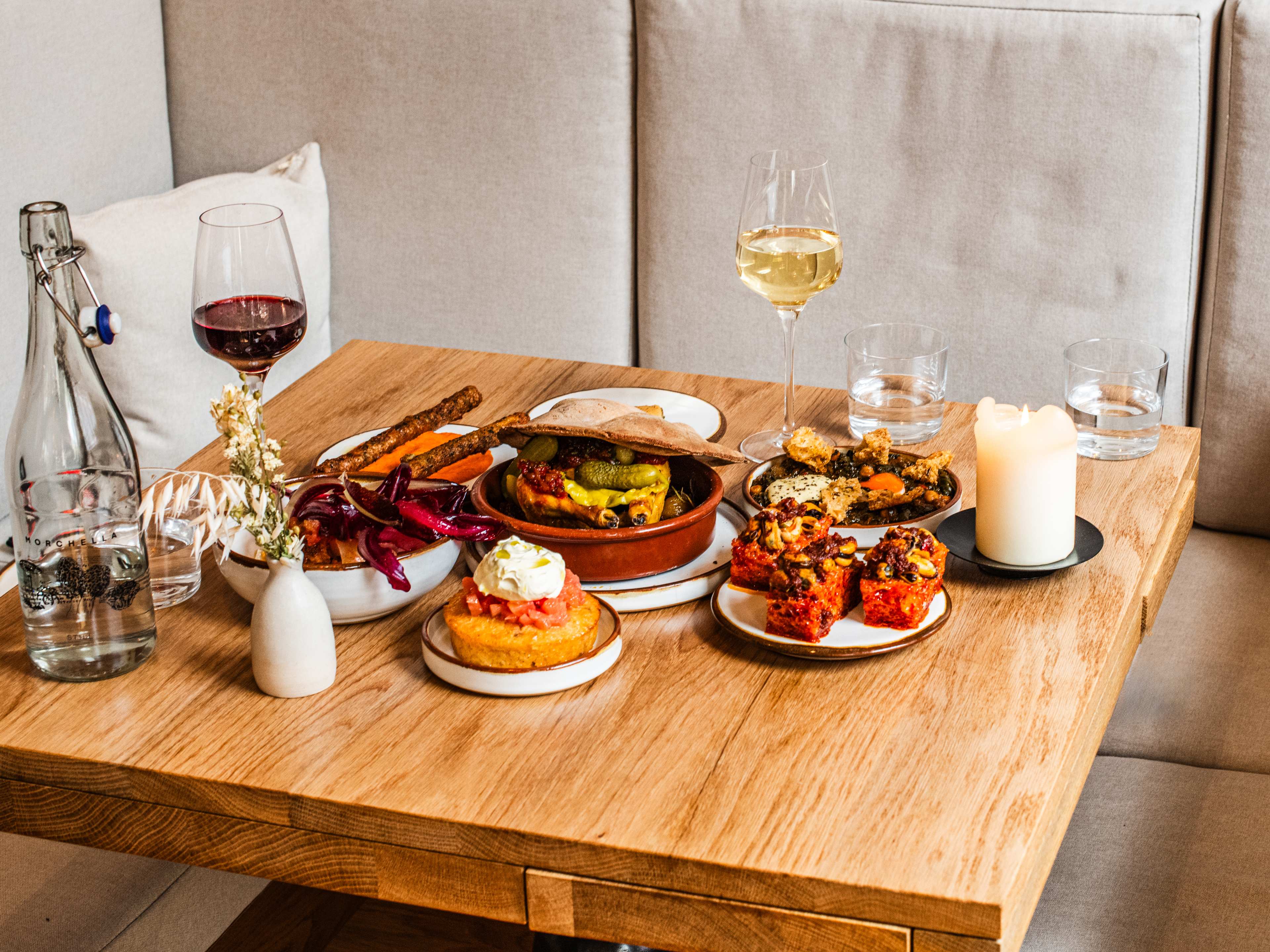 Spread of modern European dishes and glasses of wine on a wooden table, with beige booths in the background.