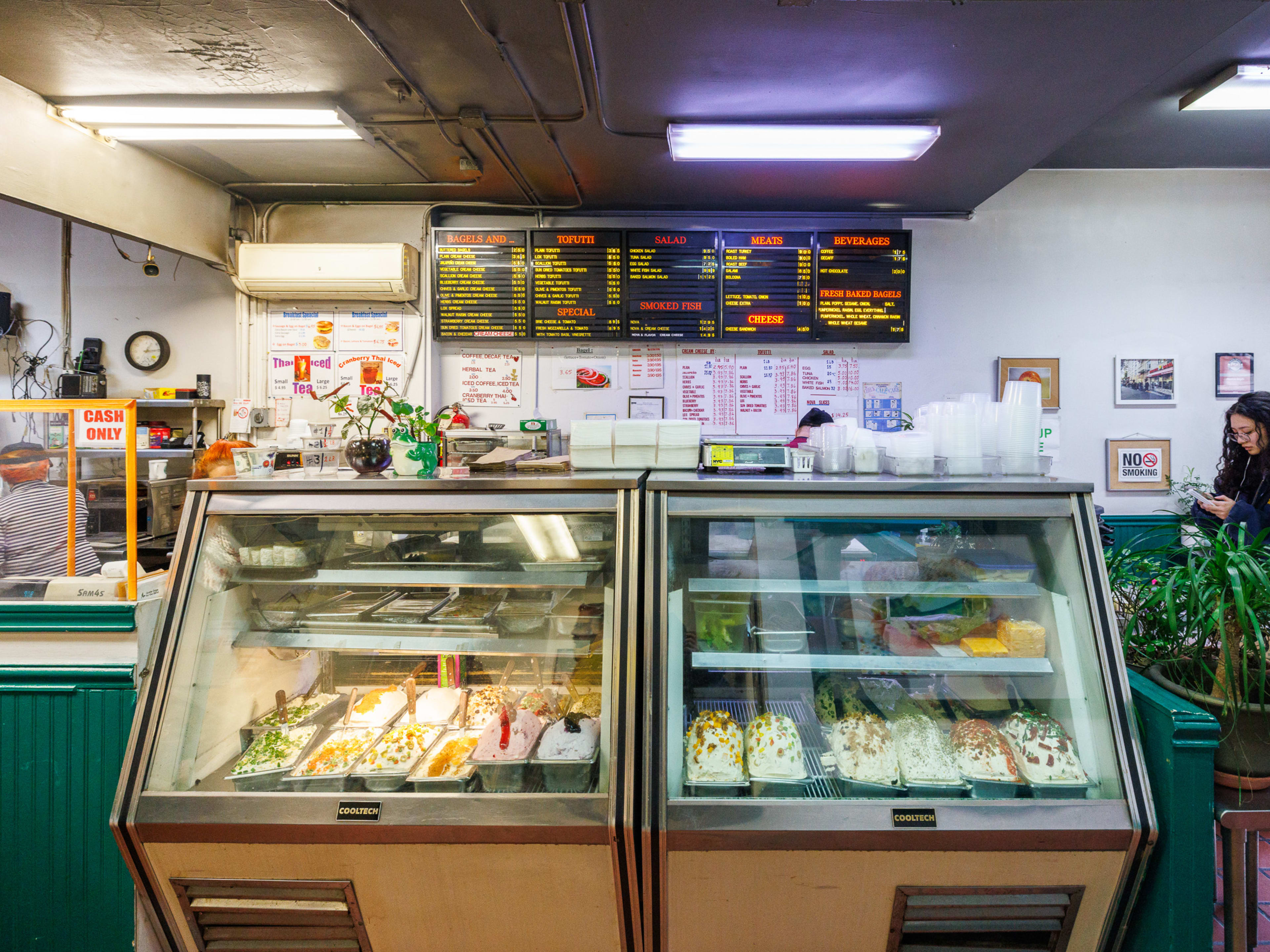 The deli case at Absolute Bagels. There is a person waiting for their order on the right and an employee working behind the counter on the left.