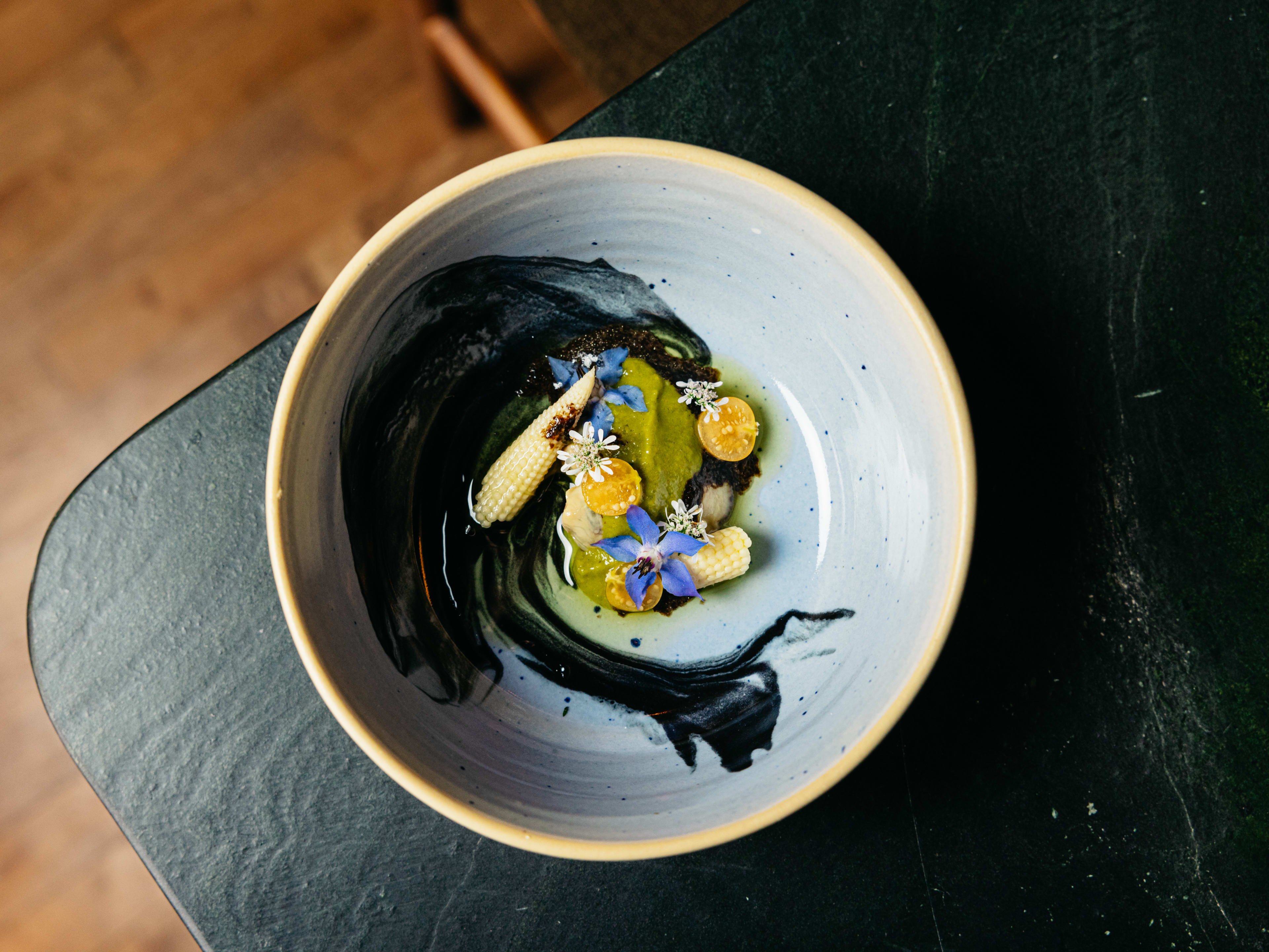 A bowl of pickled oysters, unagi sauce, baby corn, kumquats, edible flowers on the counter.