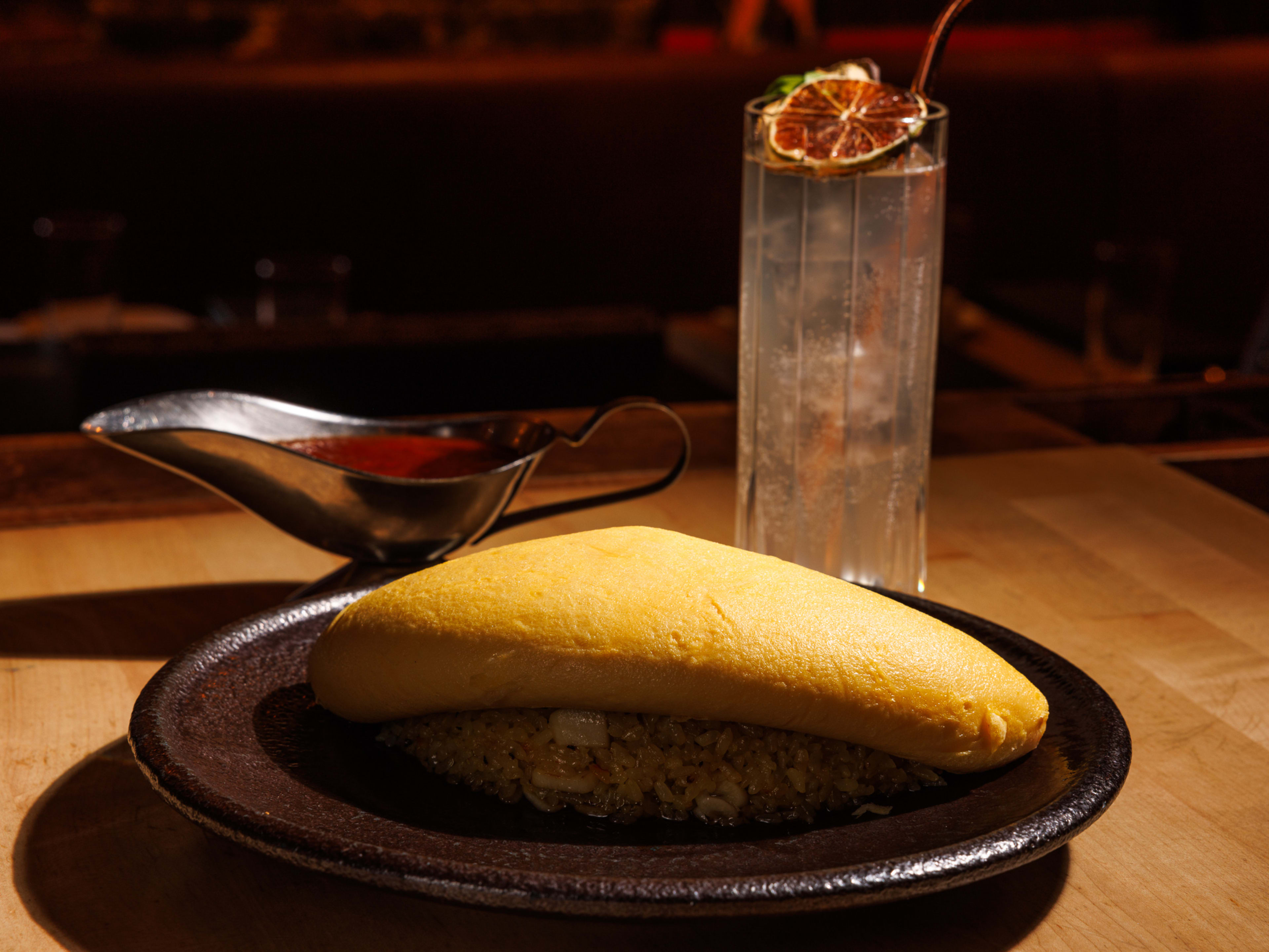 The Garlic Omu Rice still intact and served on a grey plate with a saucière and a highball cocktail on the side.