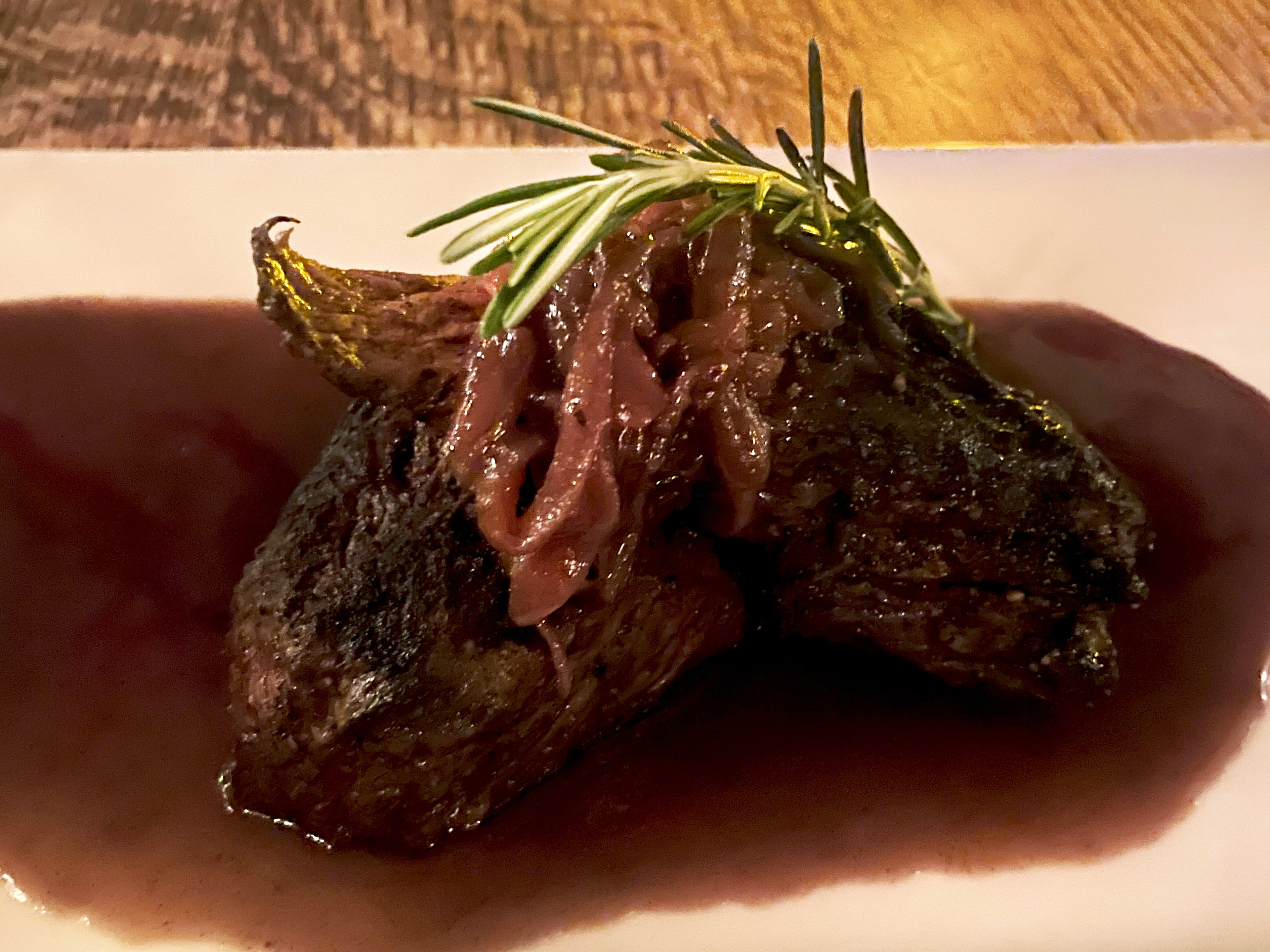 Steak in a pool of sauce on a white plate with a sprig of rosemary on top.