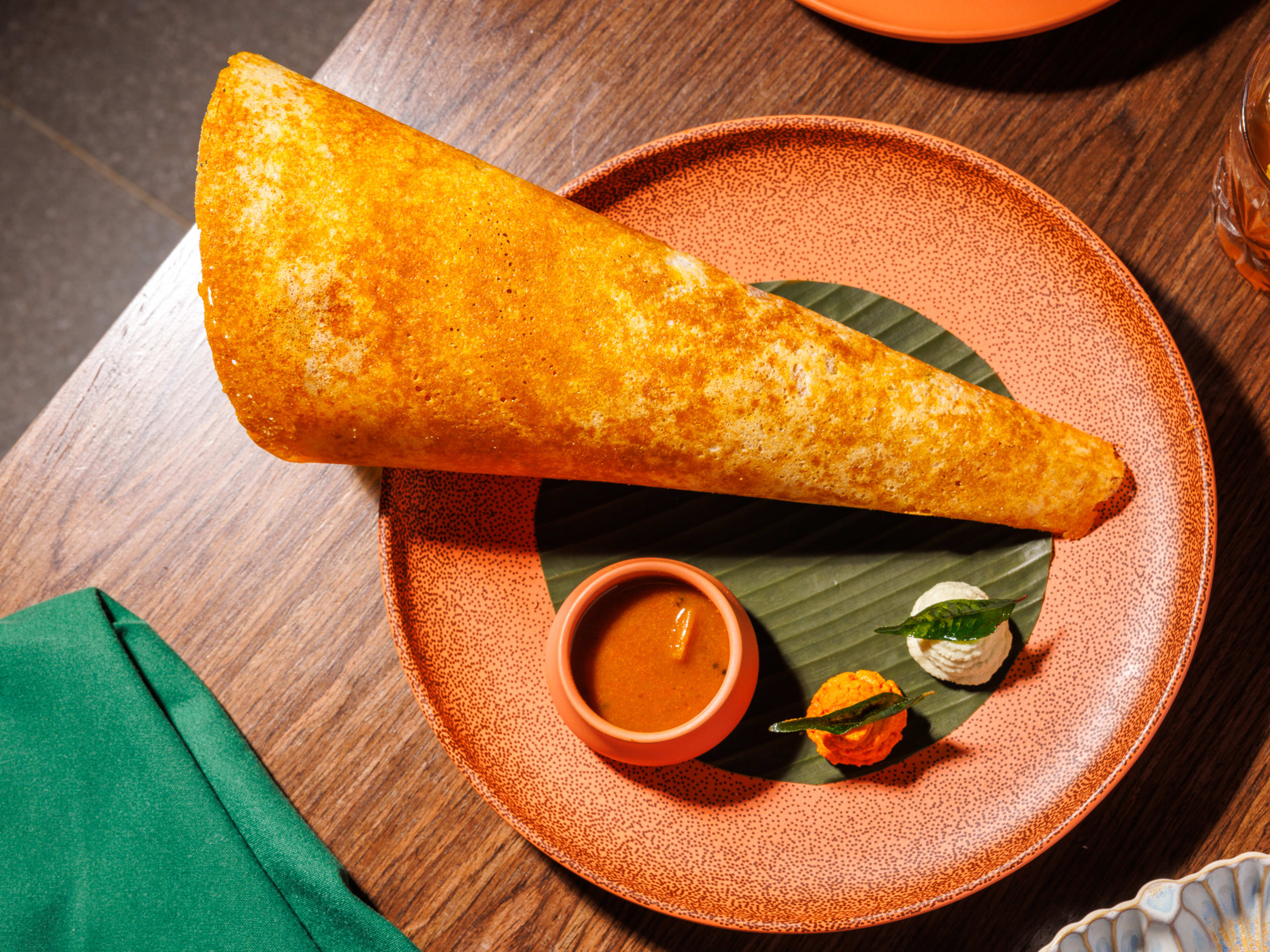 A big, golden brown dosa sitting on a plate with a few chutneys on the side.