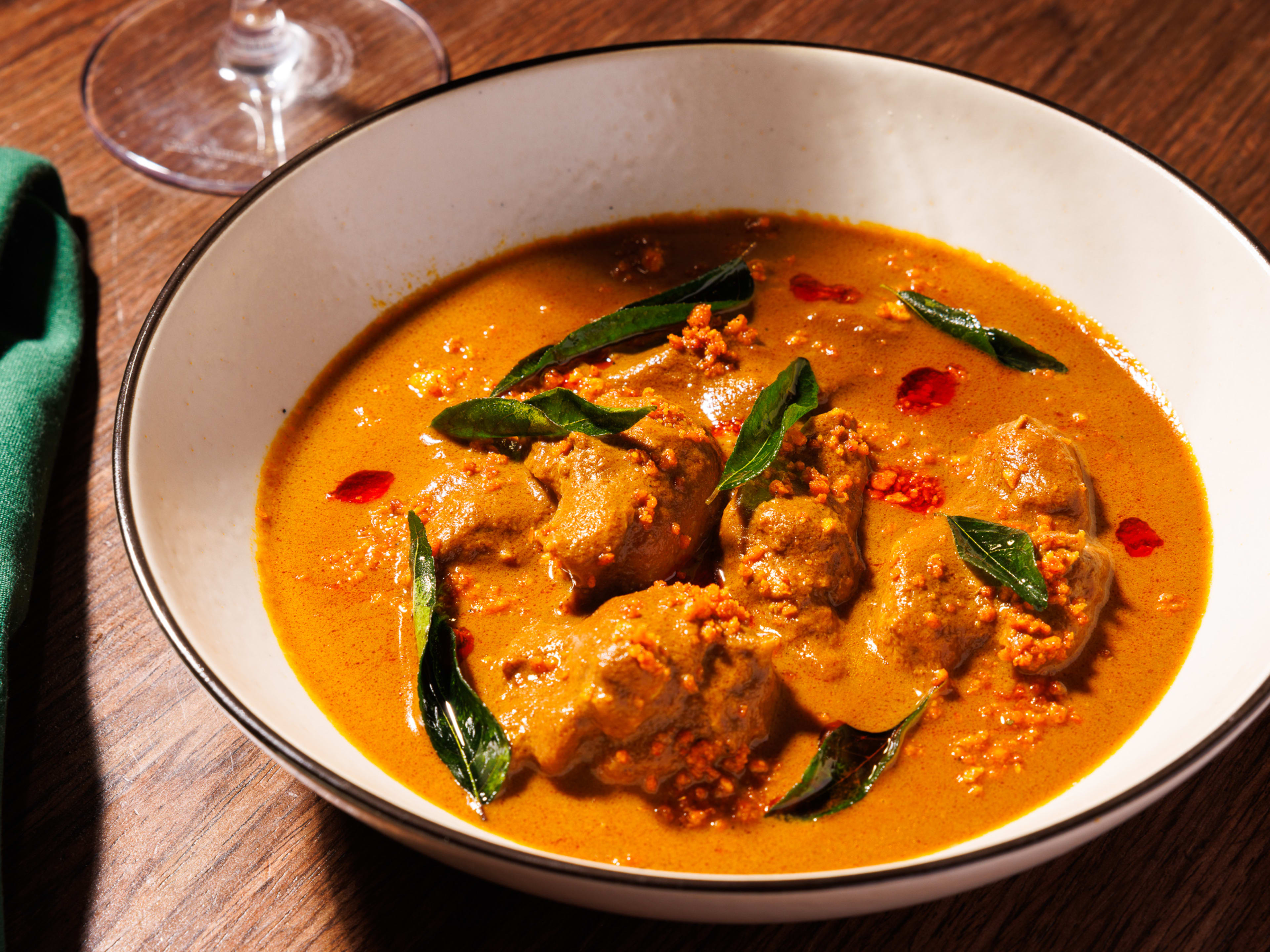 A bowl of thick, orange curry with chunks of chicken and fried curry leaves.