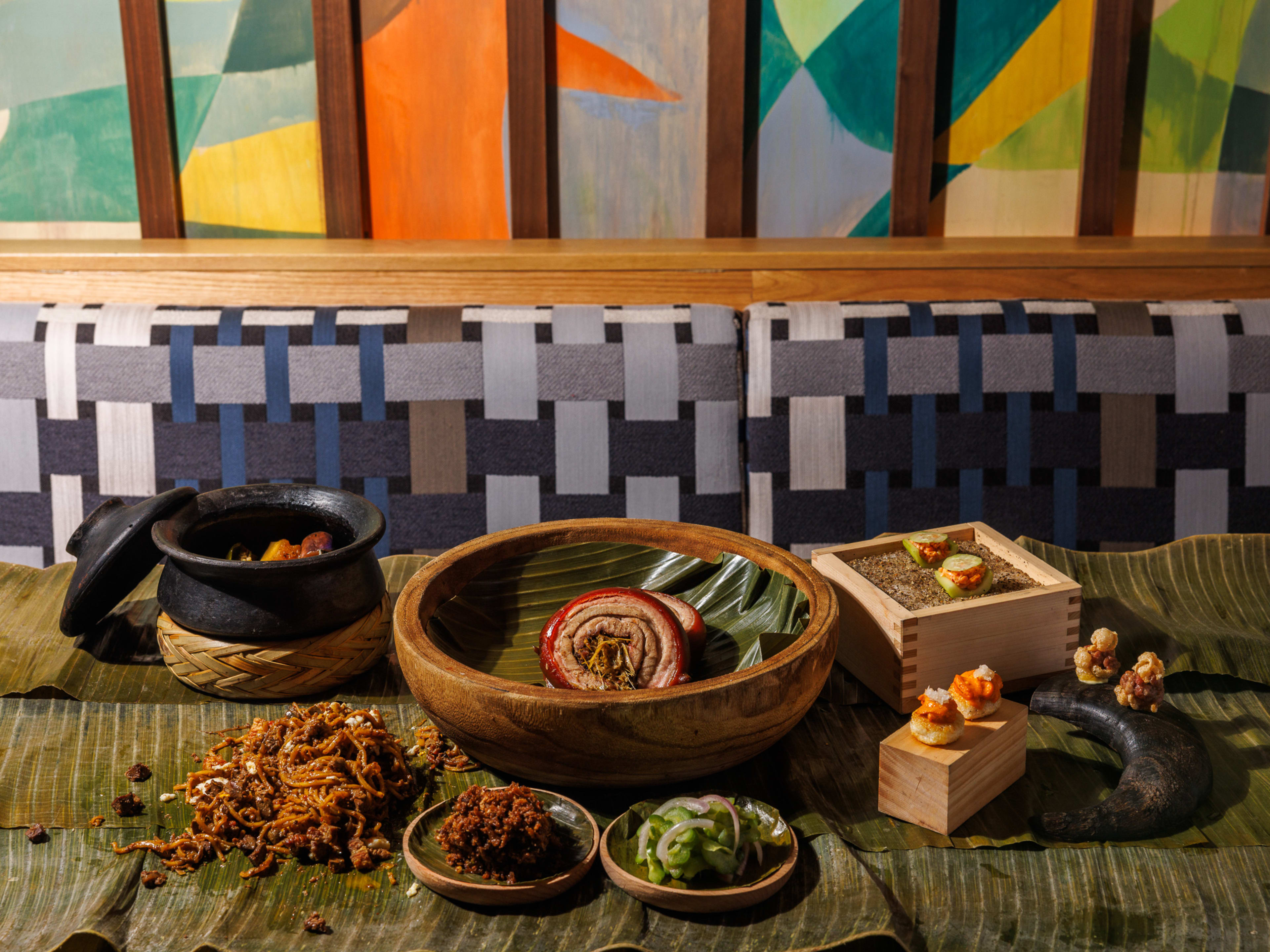 A spread of Filipino dishes on a banana leaf-covered table in front of a colorful mural.