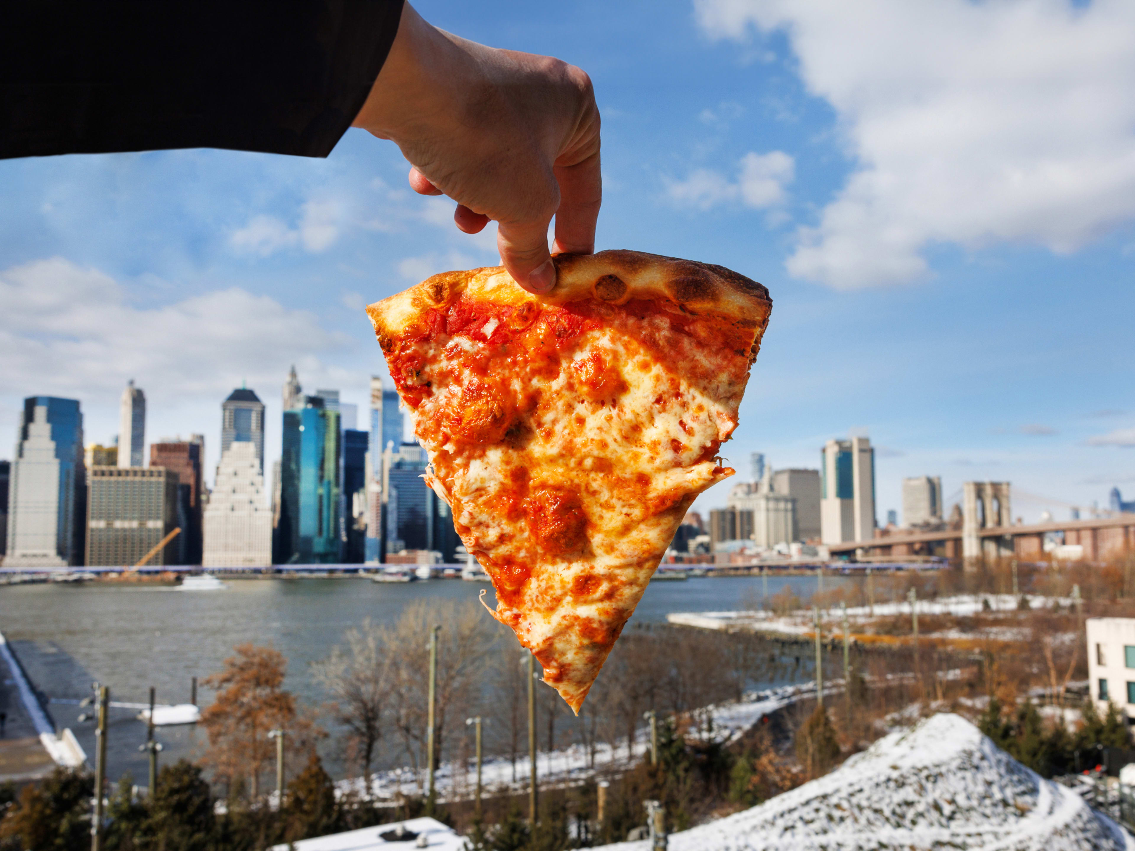 A slice of cheese pizza held up against the NYC skyline. There is a light dusting of snow on the ground.