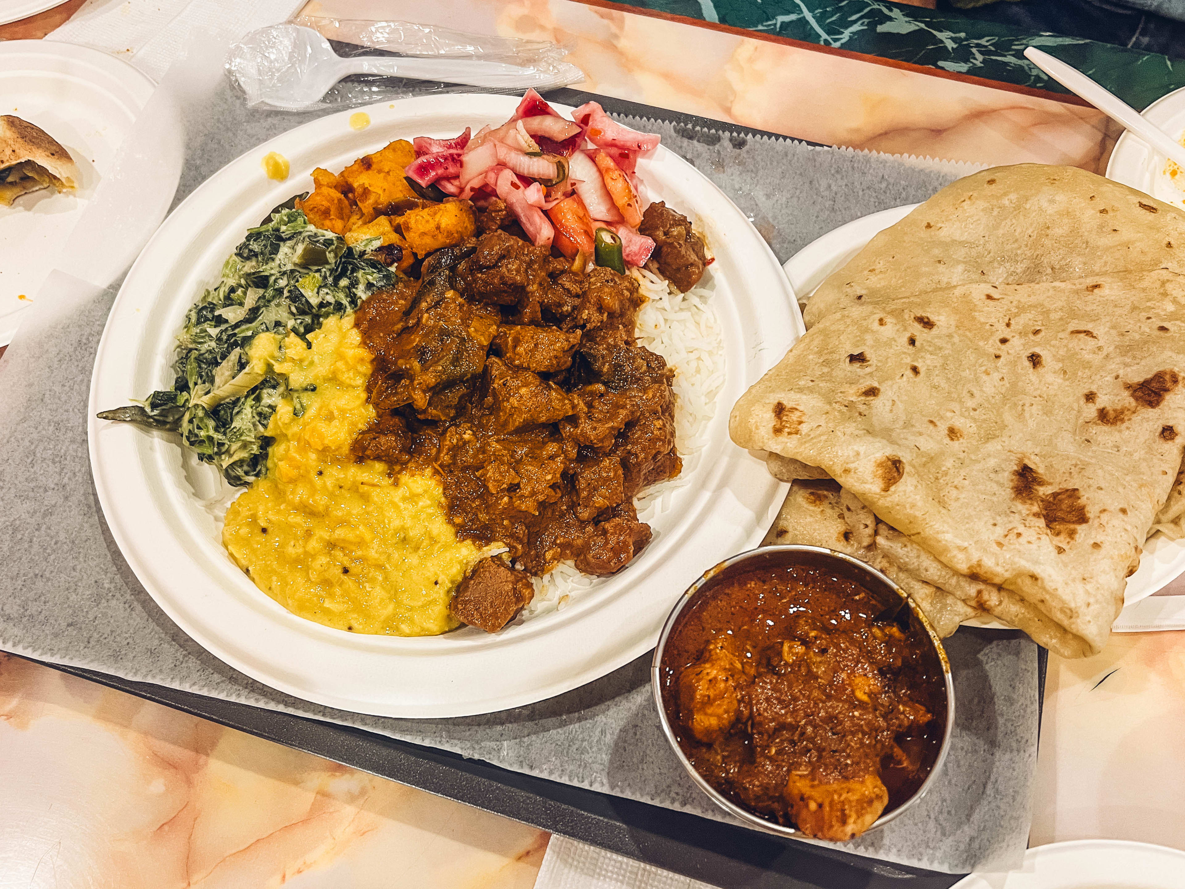 A variety of meats and vegetables on a white plate with a side of roti on a black lunch tray.