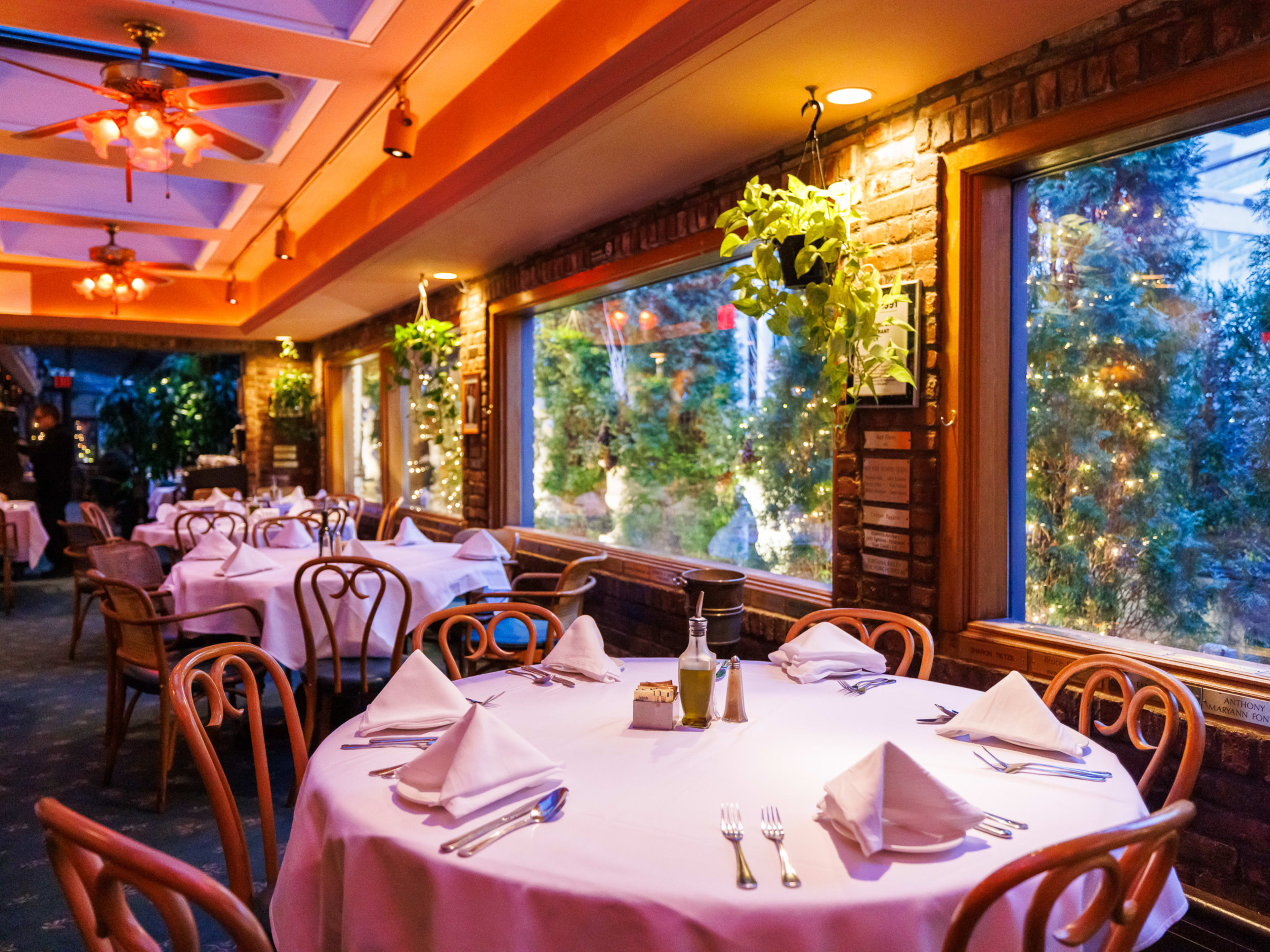 The interior of Park Side Restaurant with tables with white table clothes, planters hanging from the ceiling and large windows that look out onto a garden of foliage and sculpture.