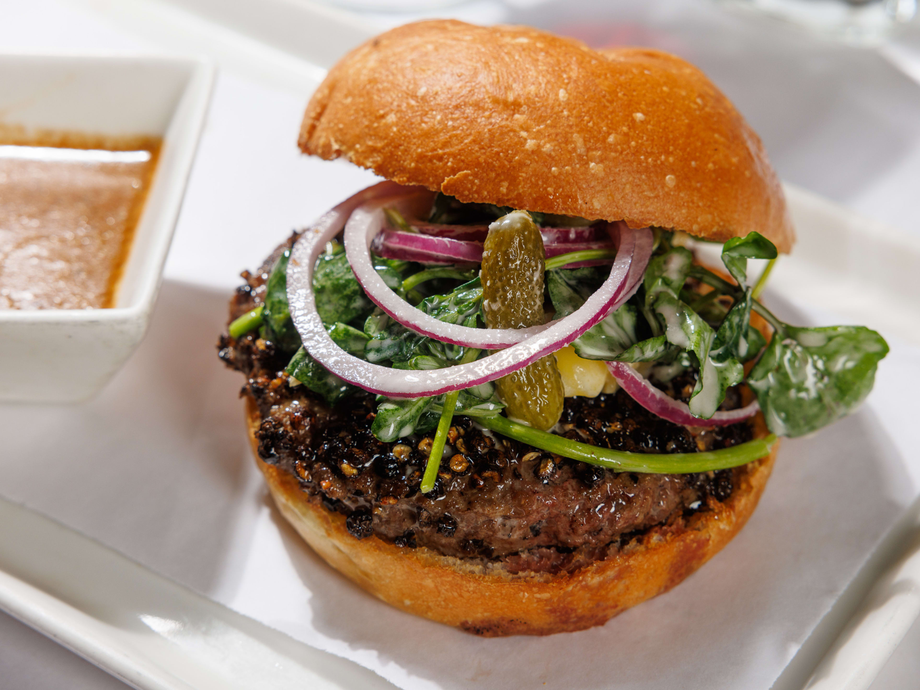 A burger with lettuce, onions, and a cornichon on a white plate with a saucer of sauce on the side.