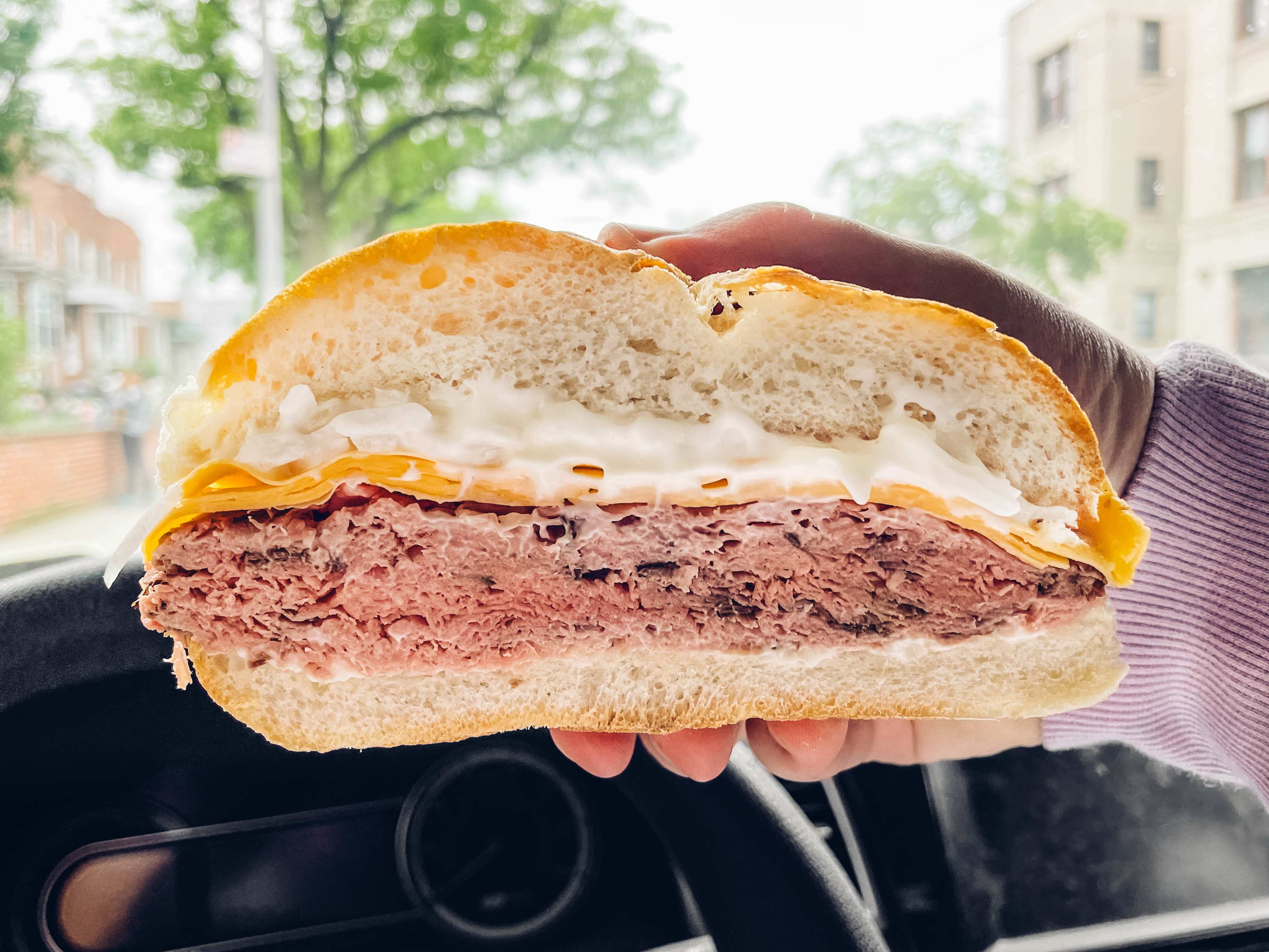 a roast beef sandwich in front of a steering wheel with cheddar, mayo, and sliced white onion