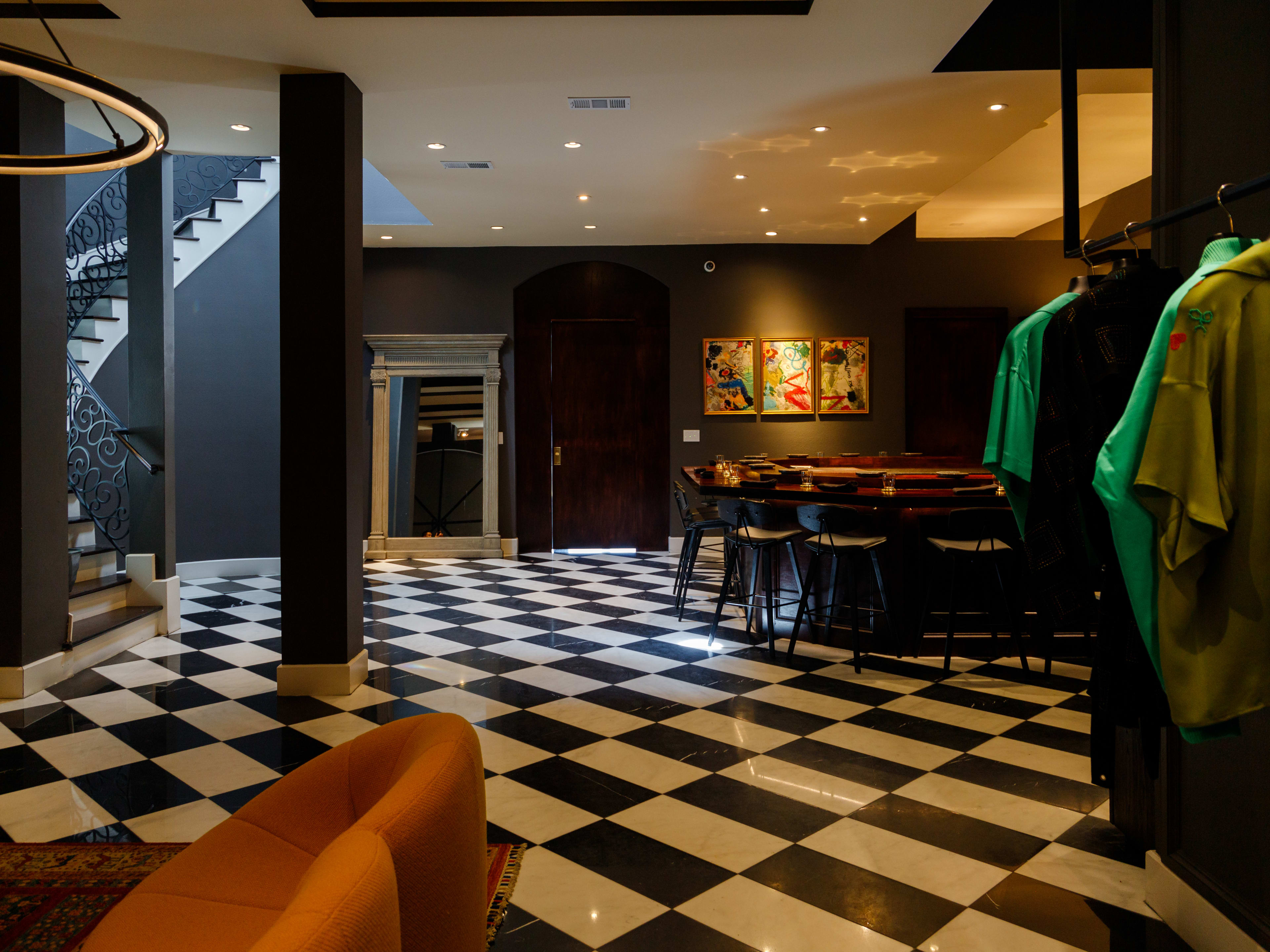 The interior of Neo HTX. There are black and white checkered floors, clothes hanging on a display to the right and a long winding staircase. The sushi counter is round and has 10 seats.