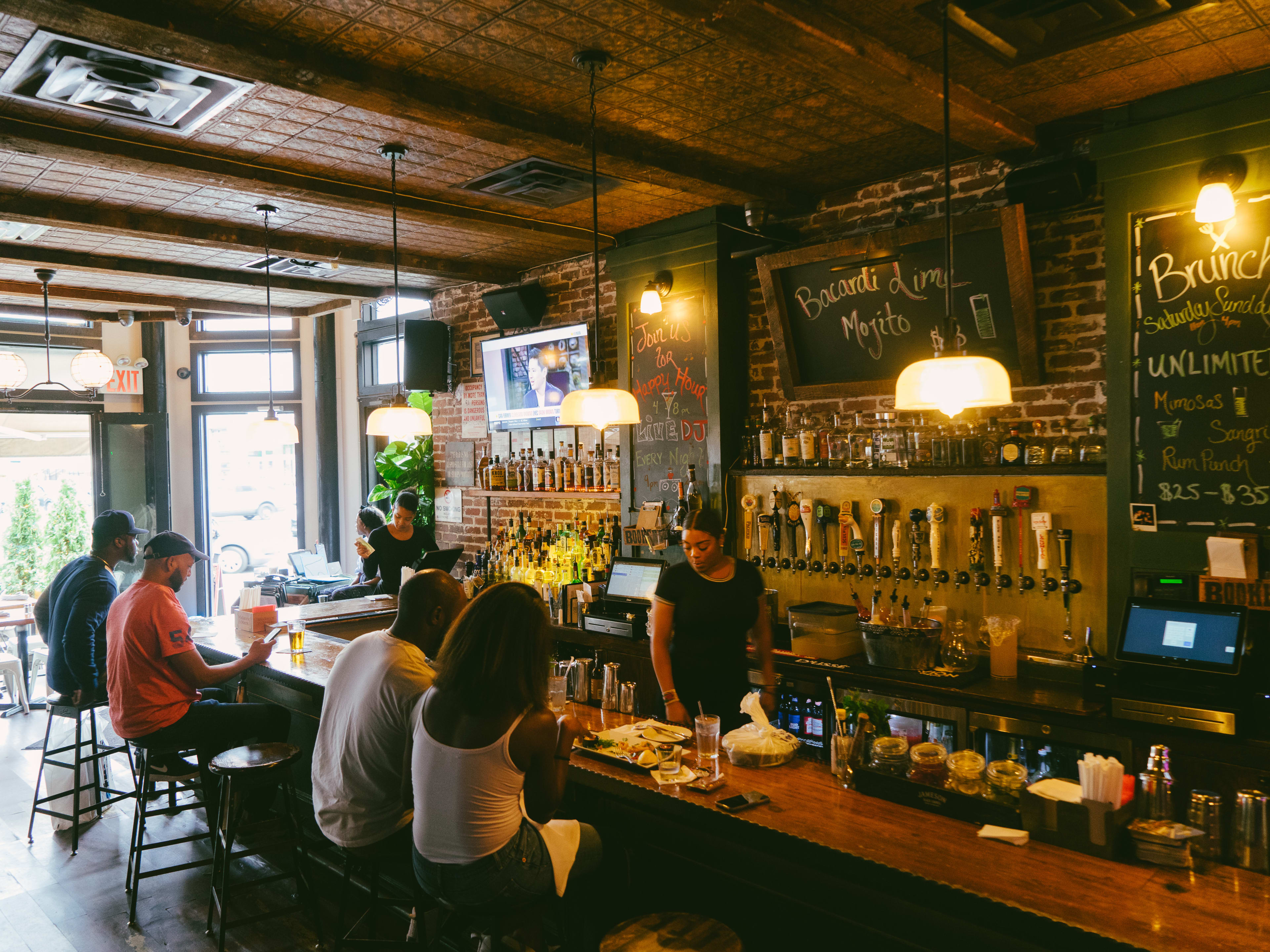 The open air interior of Corner Social. There are people sitting on stools at a wooden bar, a bartender behind the bar and one at the side.  The walls are brick and have liquor on shelves and menu items on chalkboards.
